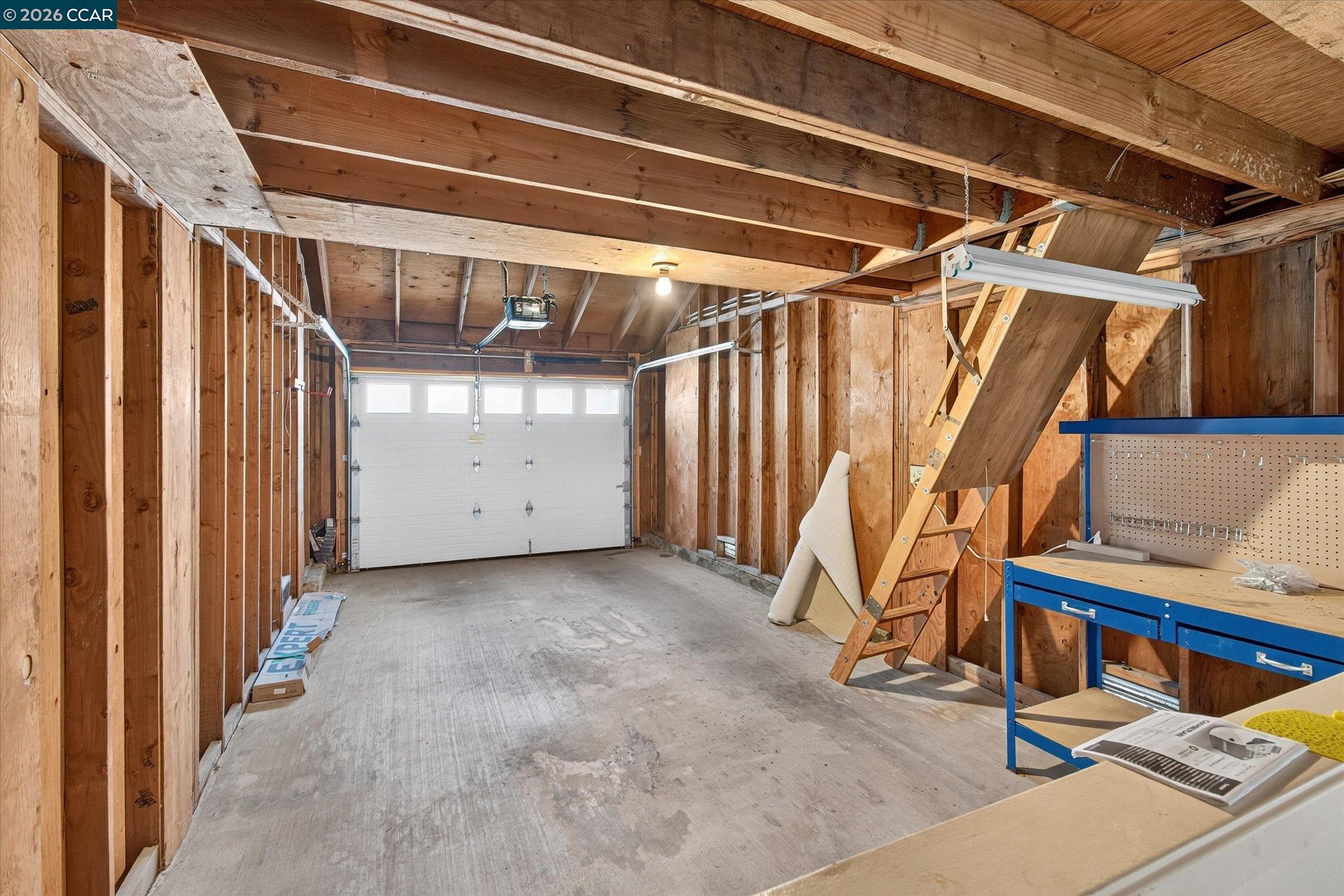 Unfinished garage with exposed framing, an attic ladder, and a blue workbench.