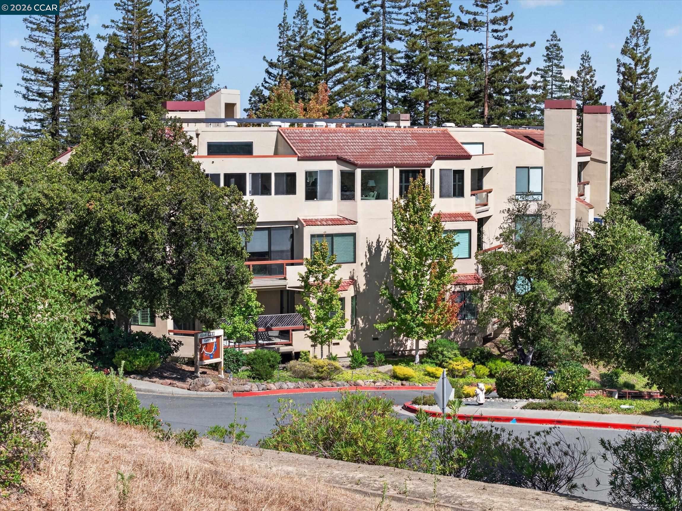 Beige multi-story residential building with red tile roofs and balconies among mature trees.