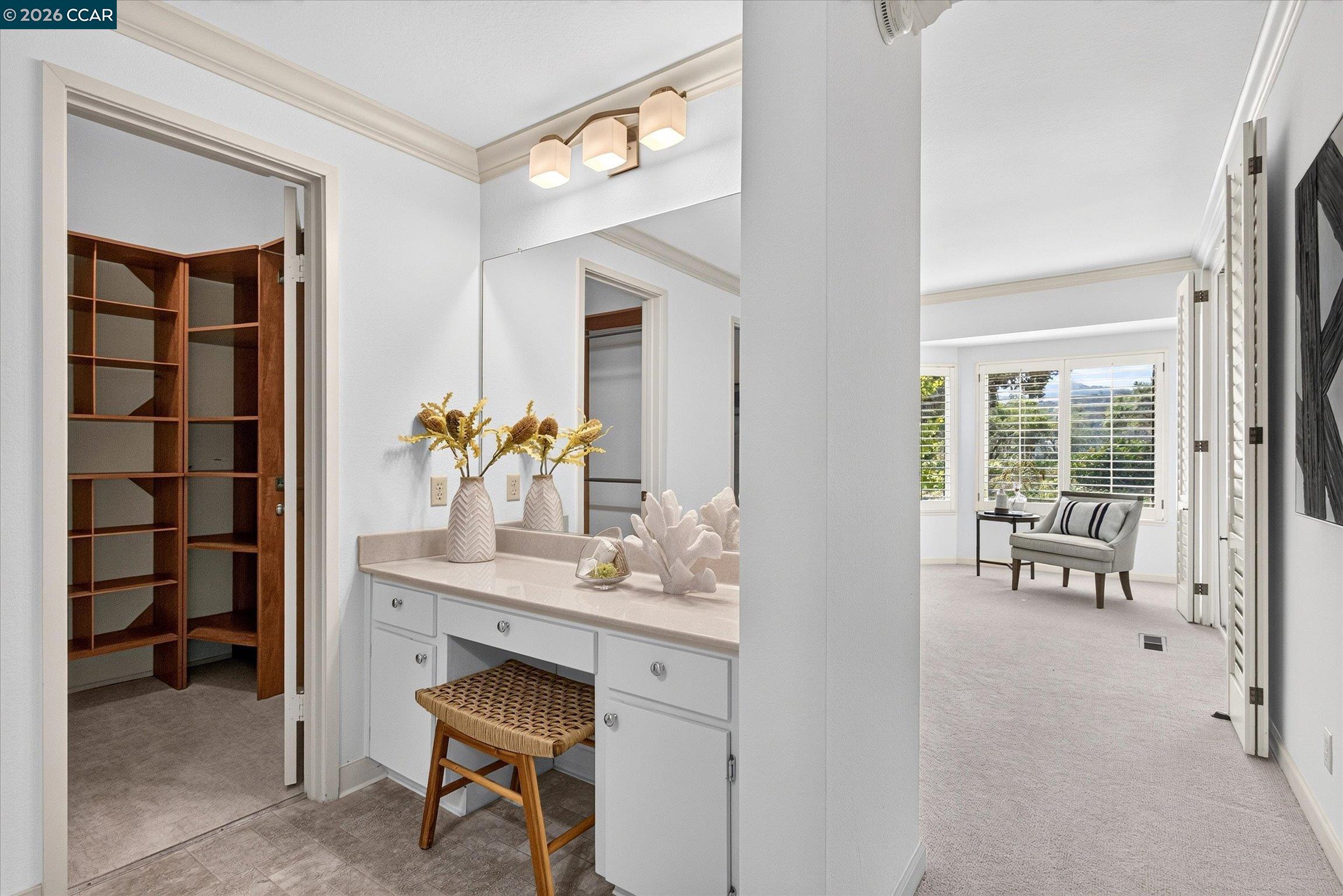 Bedroom dressing area with white vanity, wooden walk-in closet, and window-side sitting nook.