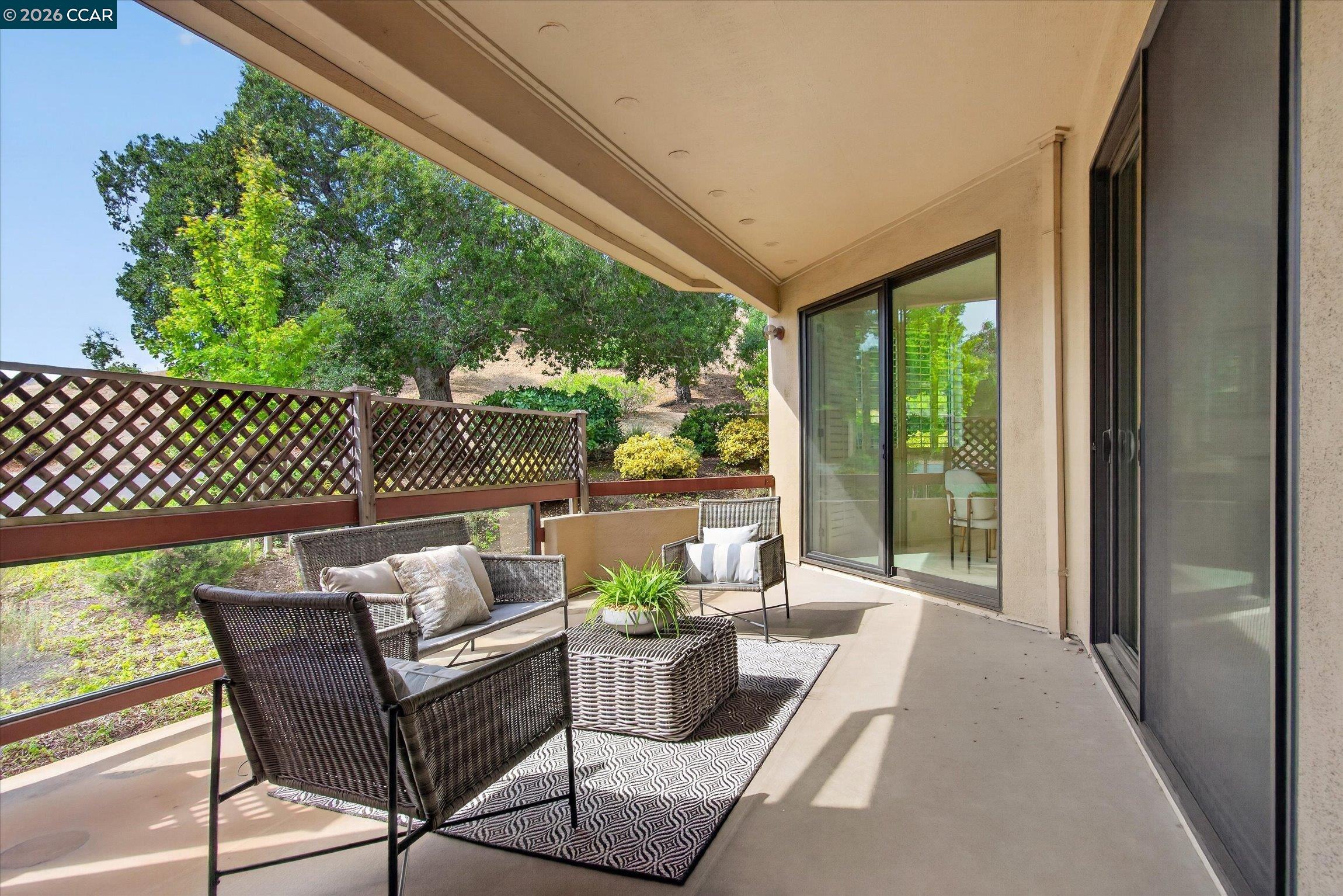 Covered patio with wicker seating and a patterned rug beside sliding glass doors, overlooking trees and shrubs.