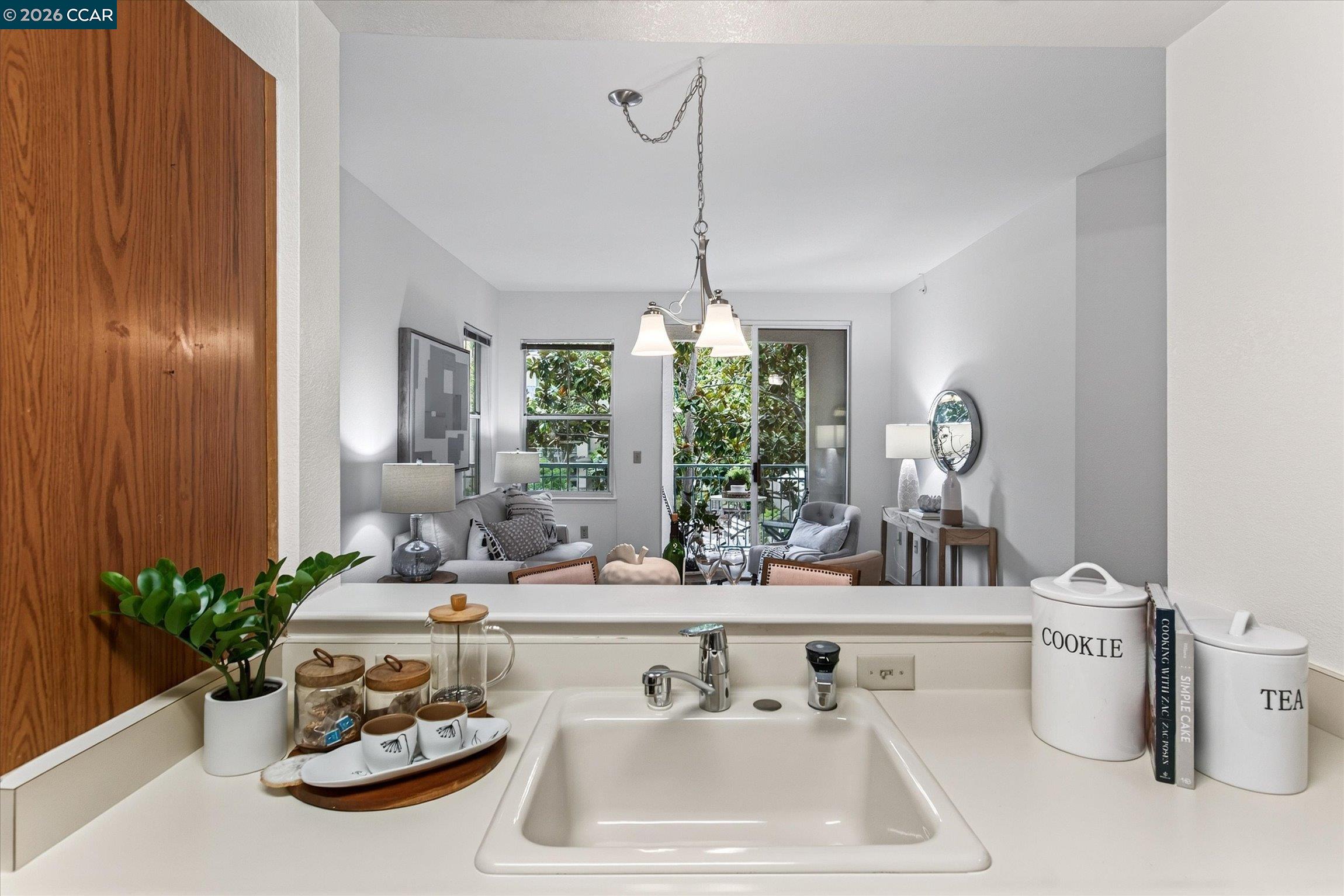 Kitchen sink and counter overlook a furnished living room with balcony doors and leafy views.