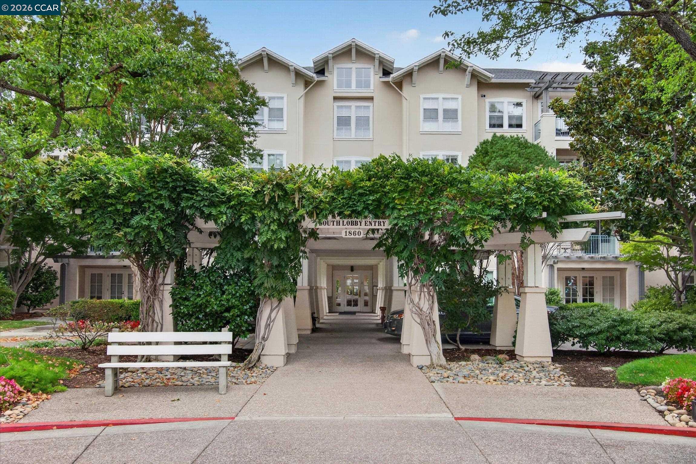 Residential building entrance framed by leafy vines, with a white bench beside the walkway.