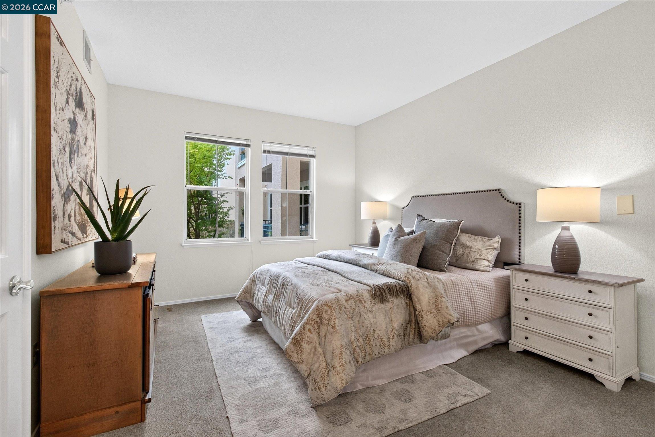 Staged bedroom with a large bed, two windows, matching bedside lamps, and a white dresser.