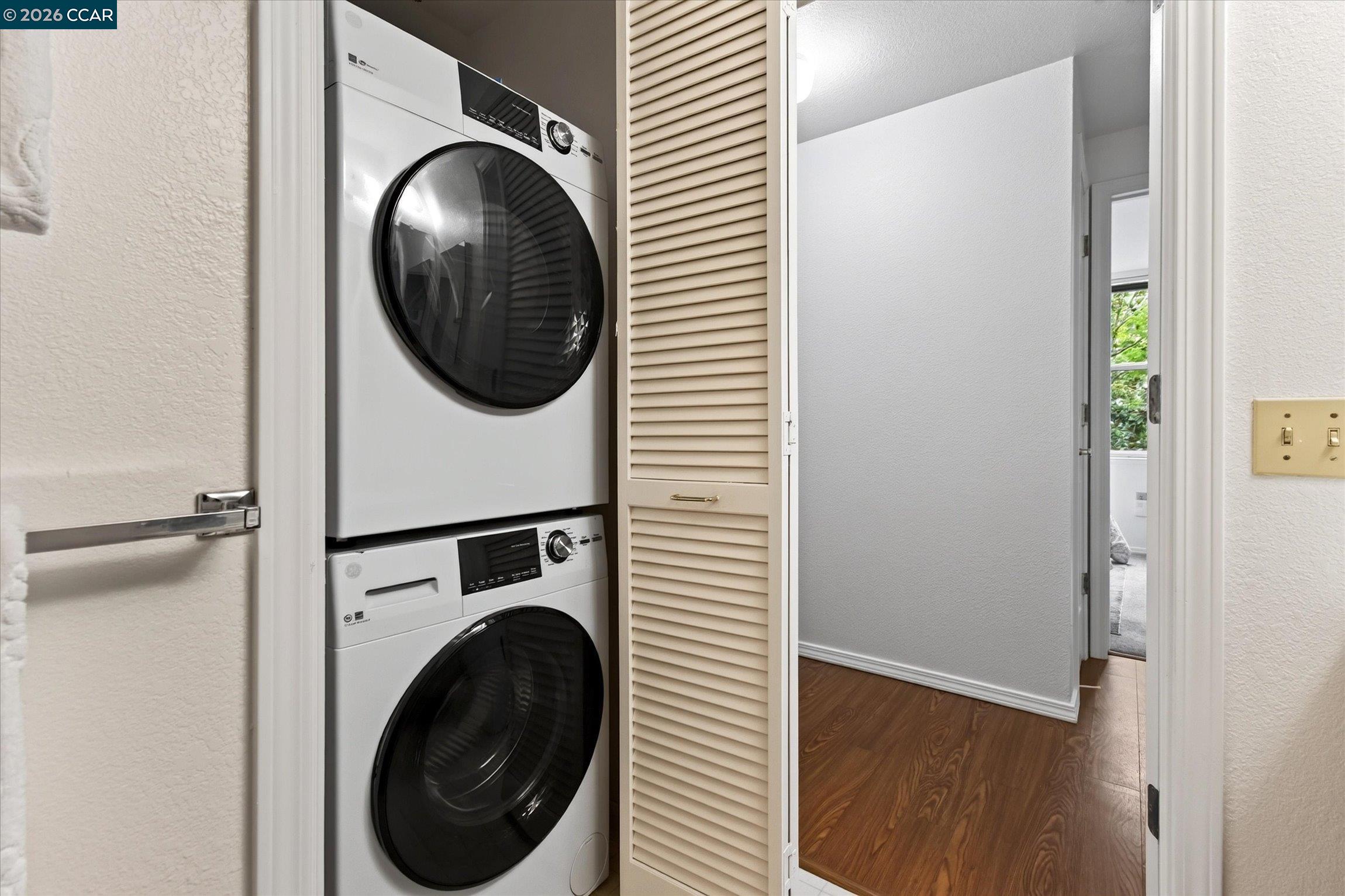 Stacked washer and dryer in a louvered laundry closet beside a wood-floored hallway.