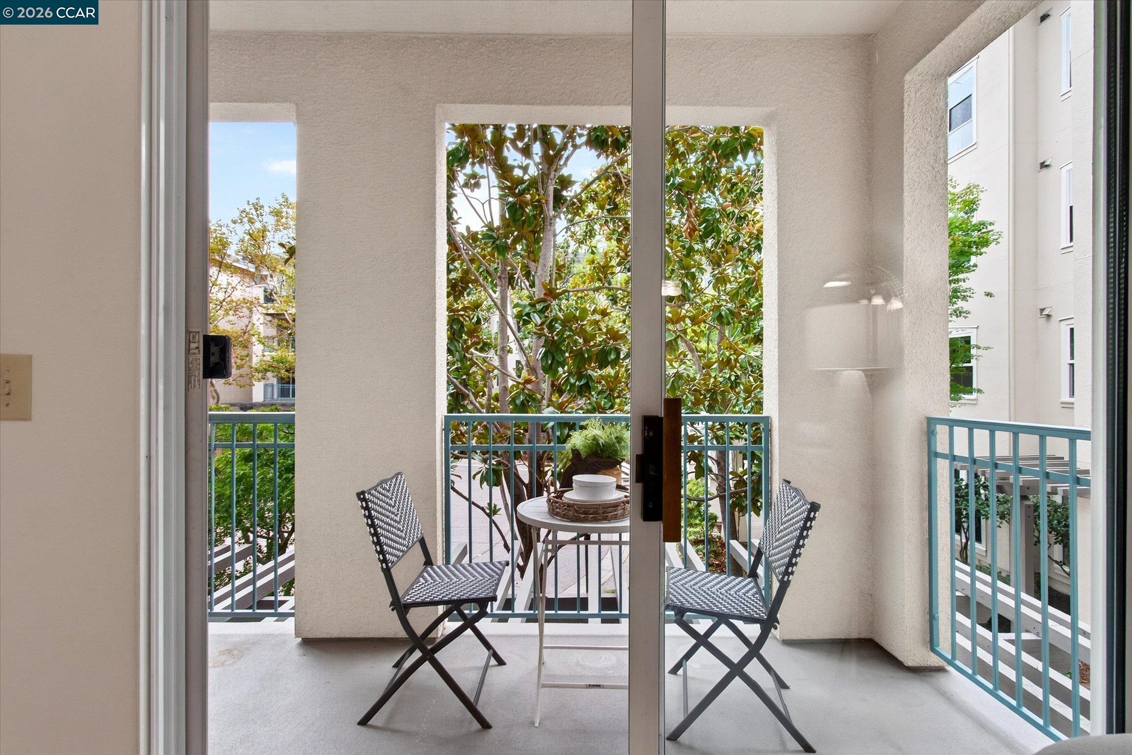 Apartment balcony with two woven chairs, a small table, blue railings, and leafy trees.