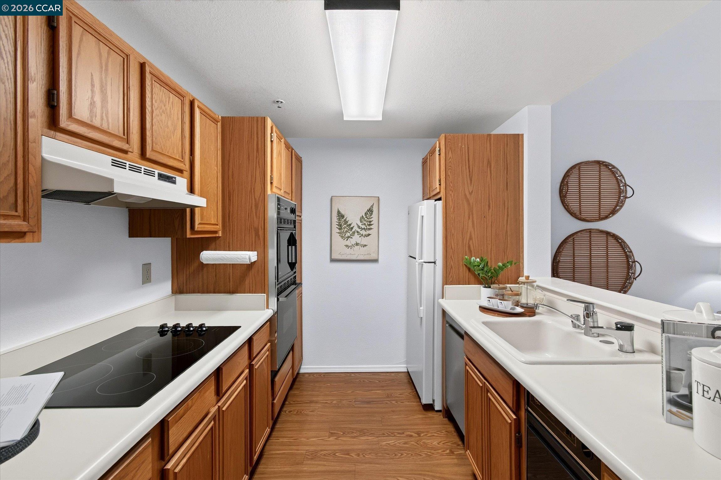 Galley kitchen with warm wood cabinets, white counters, a black cooktop, and double sink.