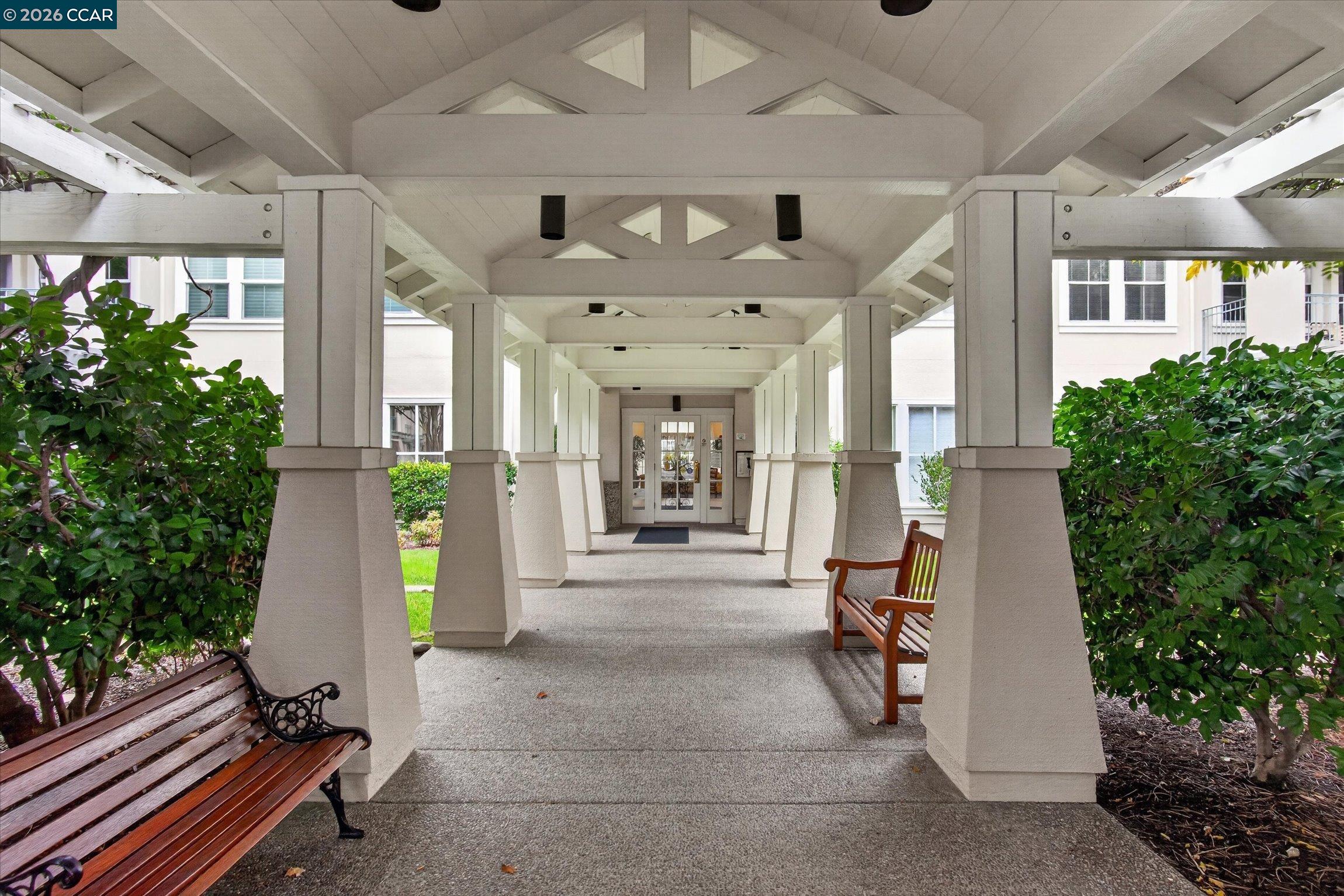 Covered walkway to the building entrance, framed by white columns, wooden benches, and greenery.