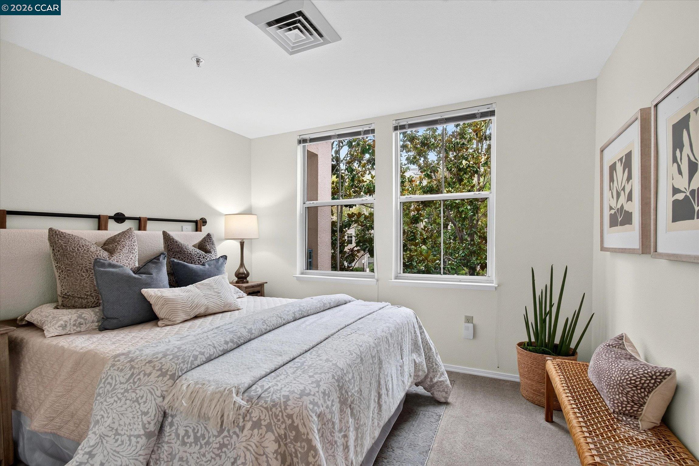 Staged bedroom with a patterned bed, two leafy-view windows, and a wicker bench.