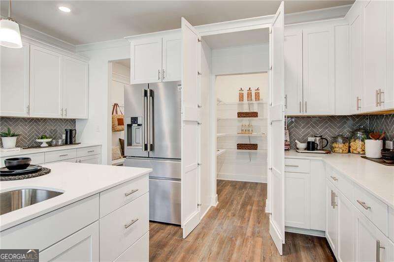 White kitchen with a large island, stainless steel refrigerator, and open pantry with wood-look flooring.