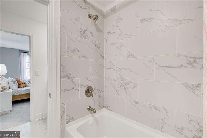 Bathroom with a white tub, chrome fixtures, and marble-look tile beside a doorway to a bedroom.