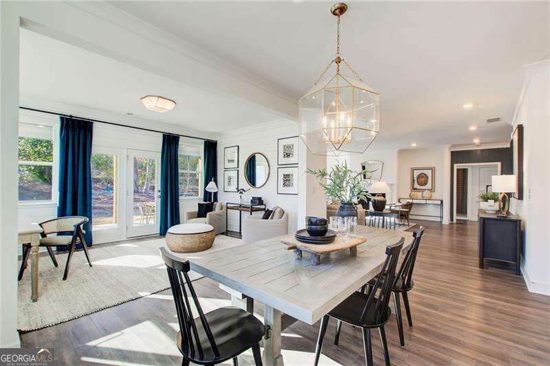 Open-plan dining and living area with a large table, geometric pendant chandelier, and bright window-side seating.