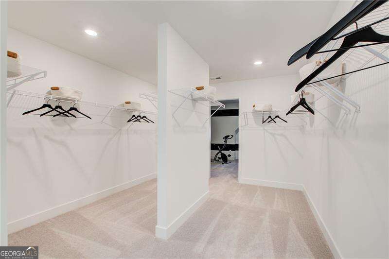 Walk-in closet with wire shelving, hanging rods, beige carpet, and an exercise bike visible through the doorway.