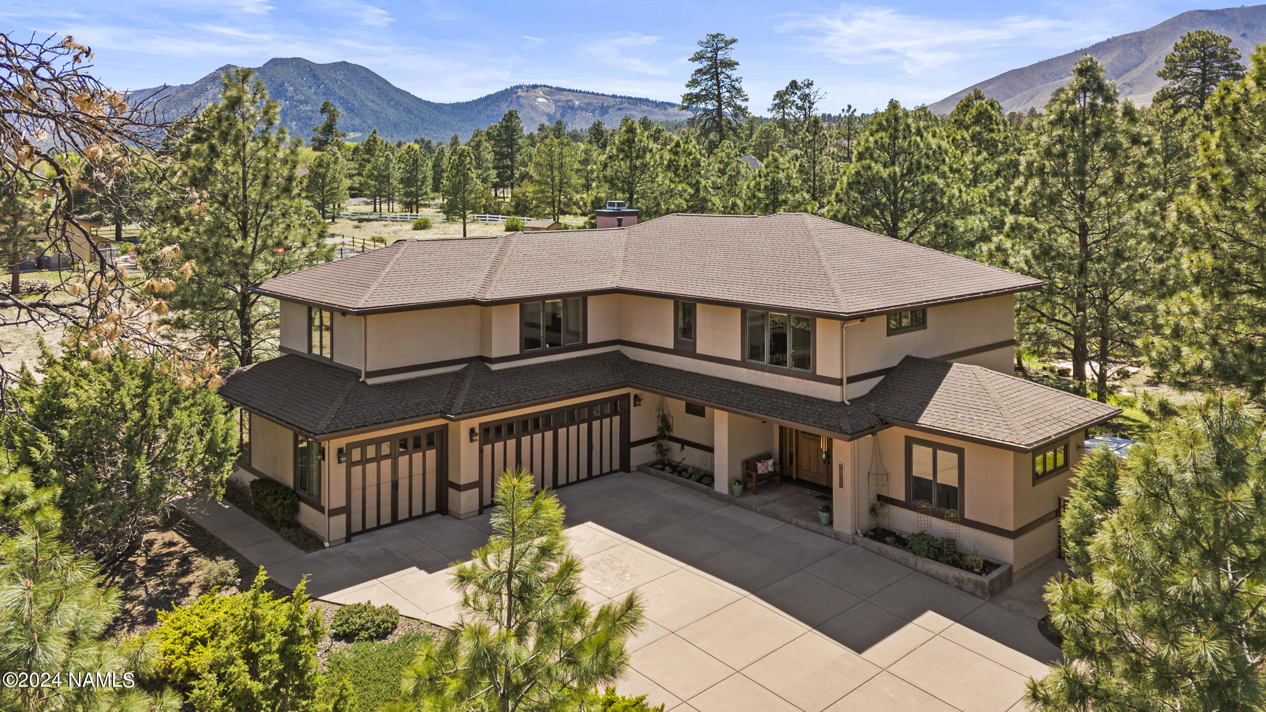 Two-story beige home with multiple garage doors, surrounded by pine trees and mountain views.