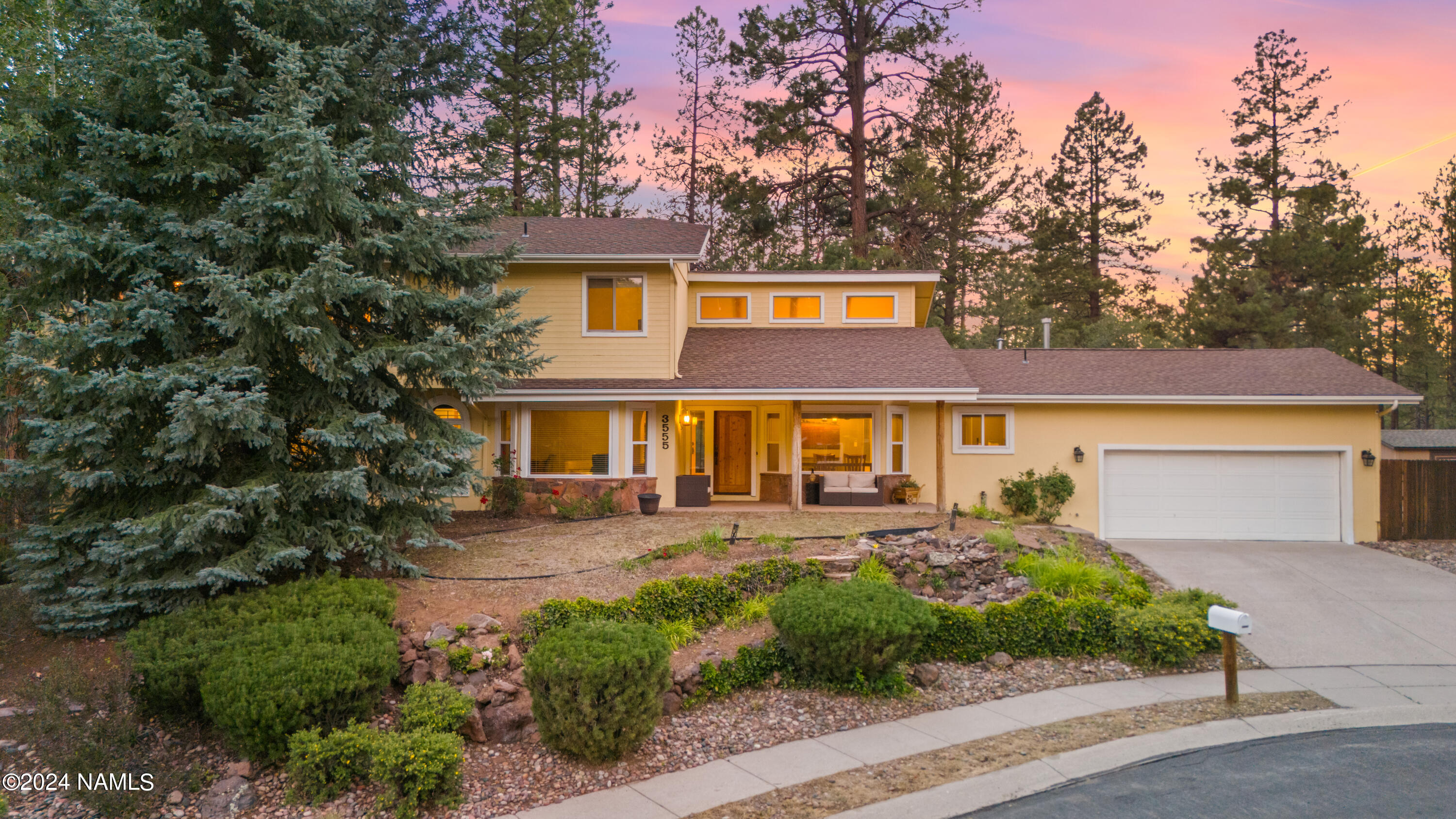 Yellow two-story home with an attached garage, evergreen trees, and warm lights glowing at dusk.