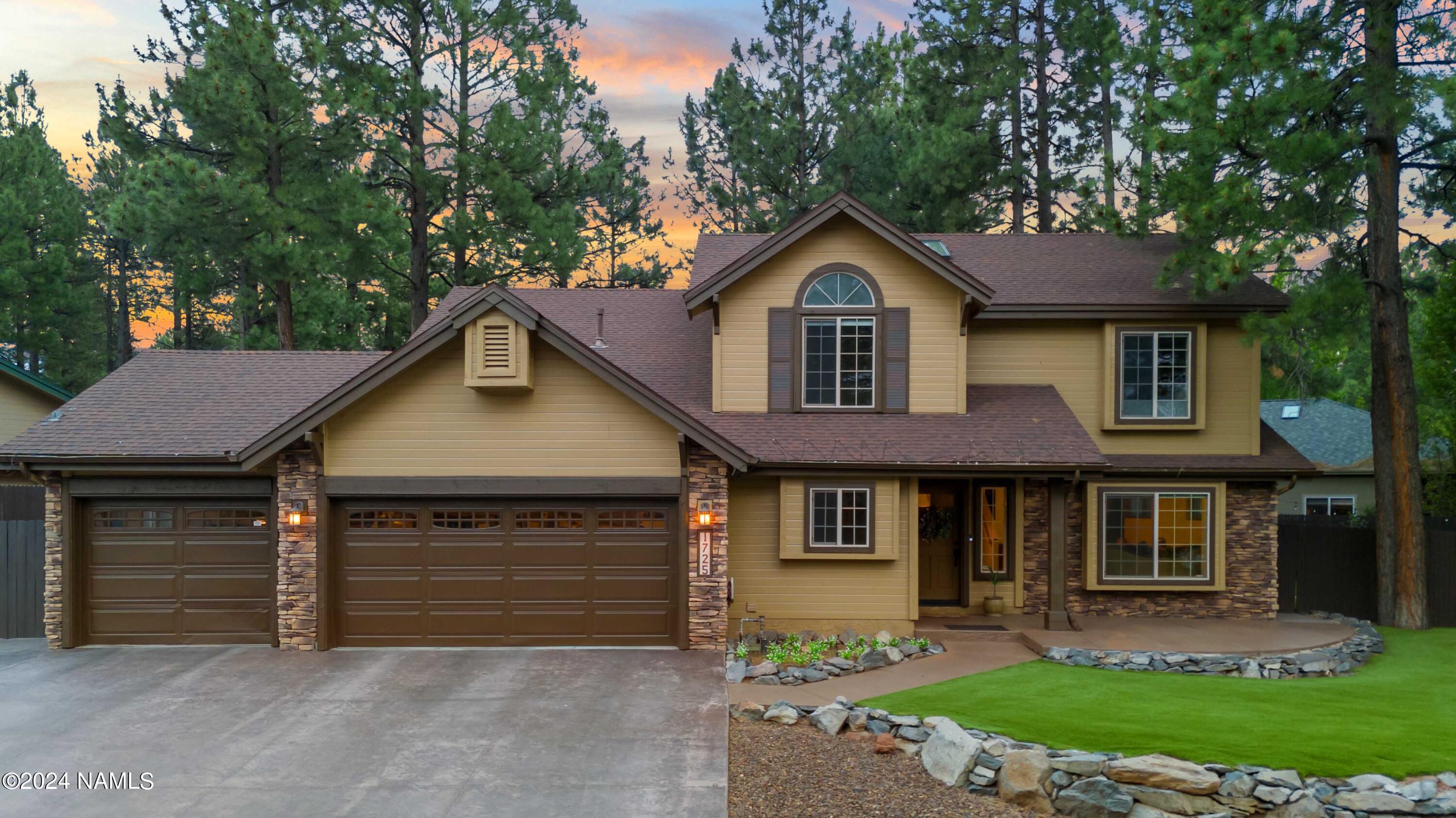 Two-story tan home with stone accents, two garage doors, and a landscaped lawn among pine trees at dusk.
