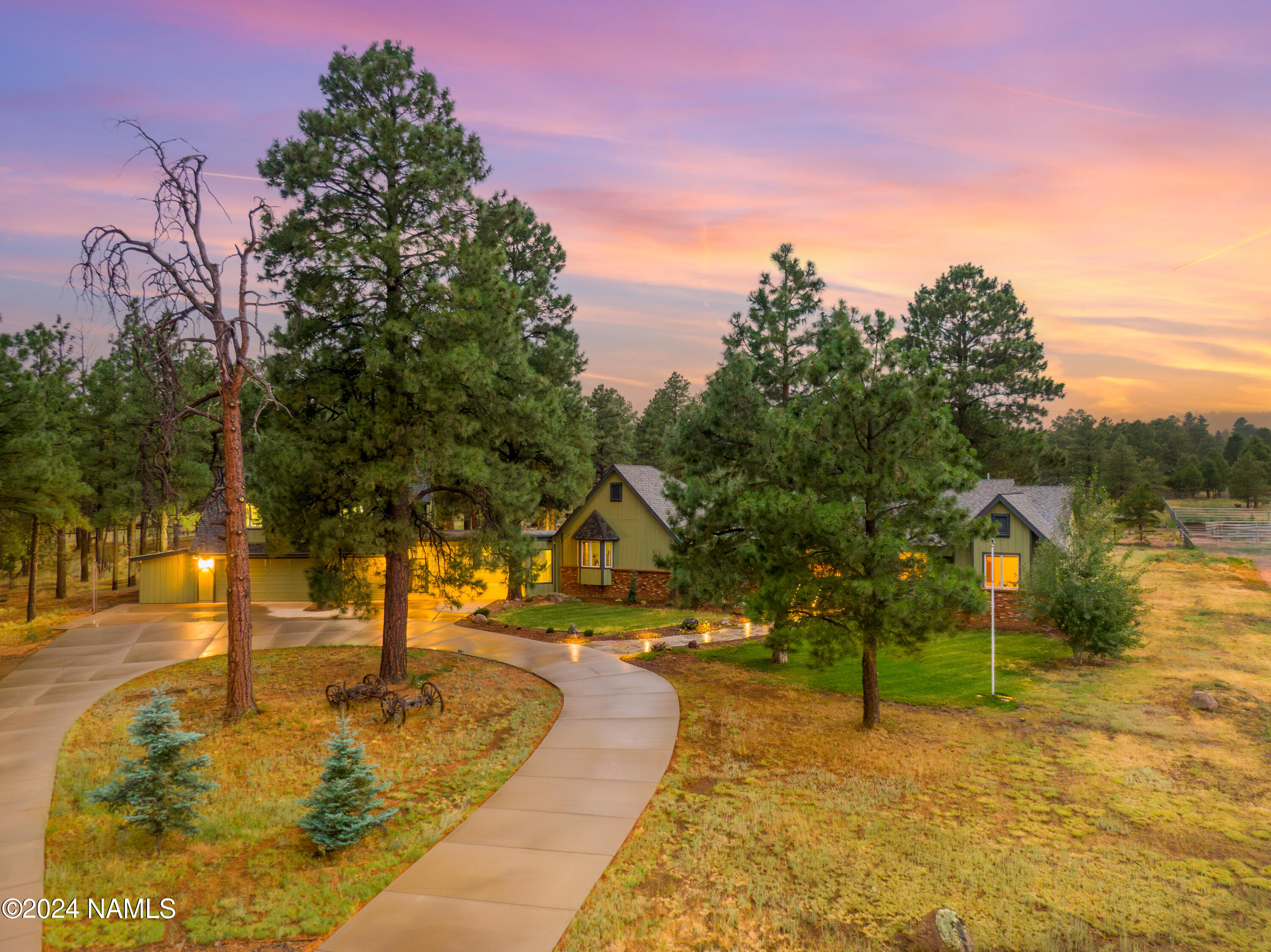 Home among tall pines at dusk, with a curved walkway and warmly lit windows.