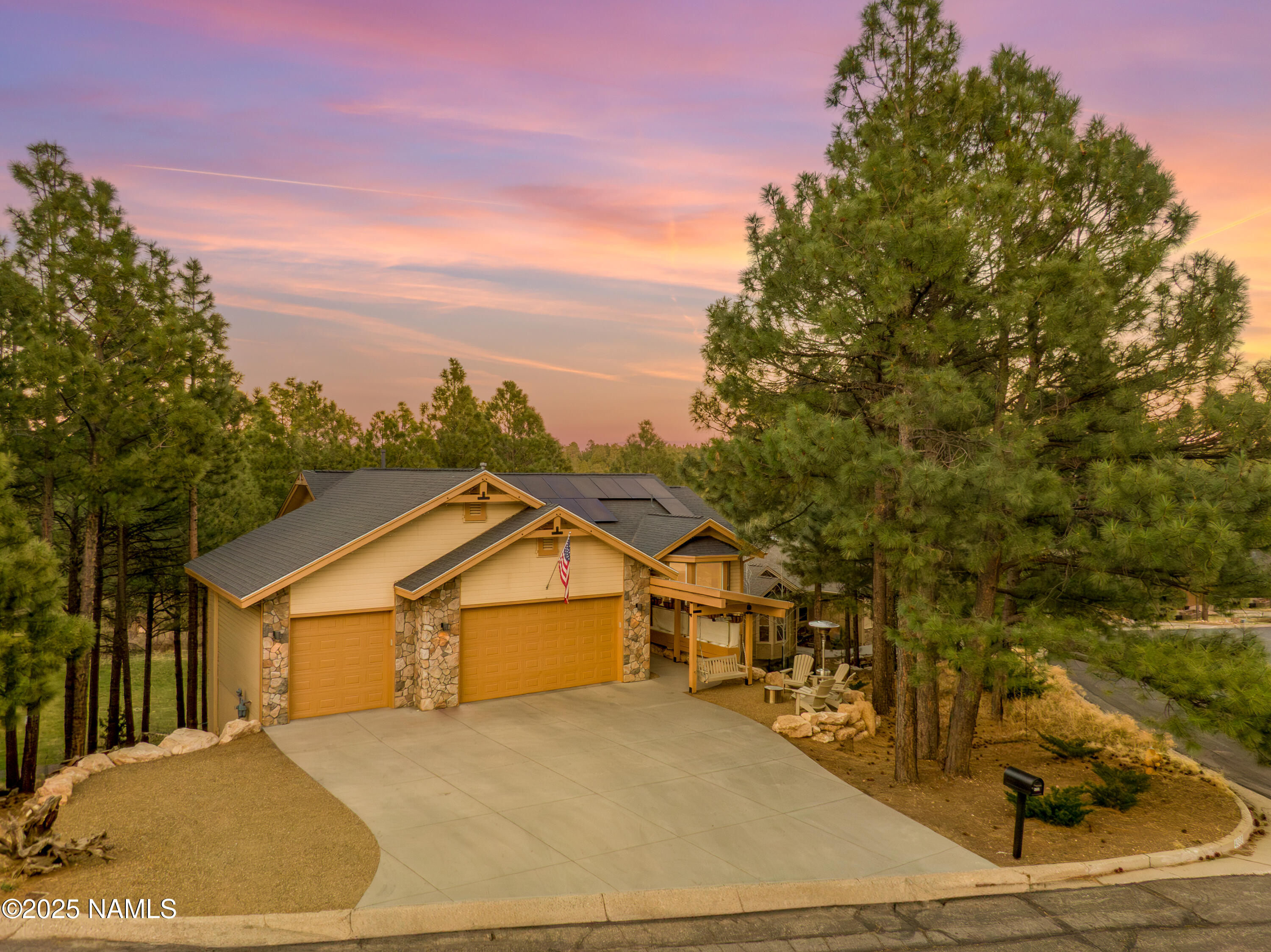 Tan-and-stone home with a broad driveway and pine trees beneath a colorful sunset sky.