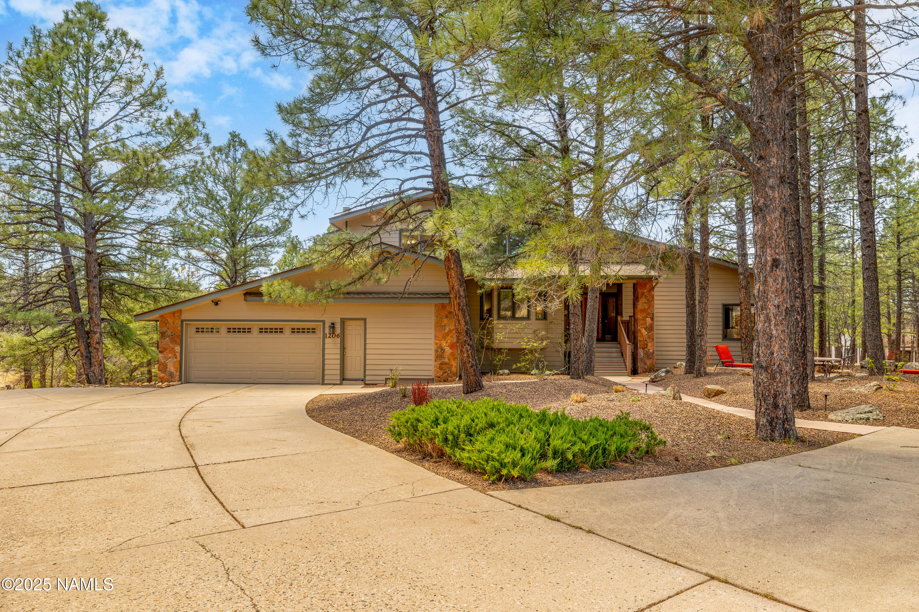 Tan home with stone accents and a two-car garage nestled among tall pines beside a curved driveway.