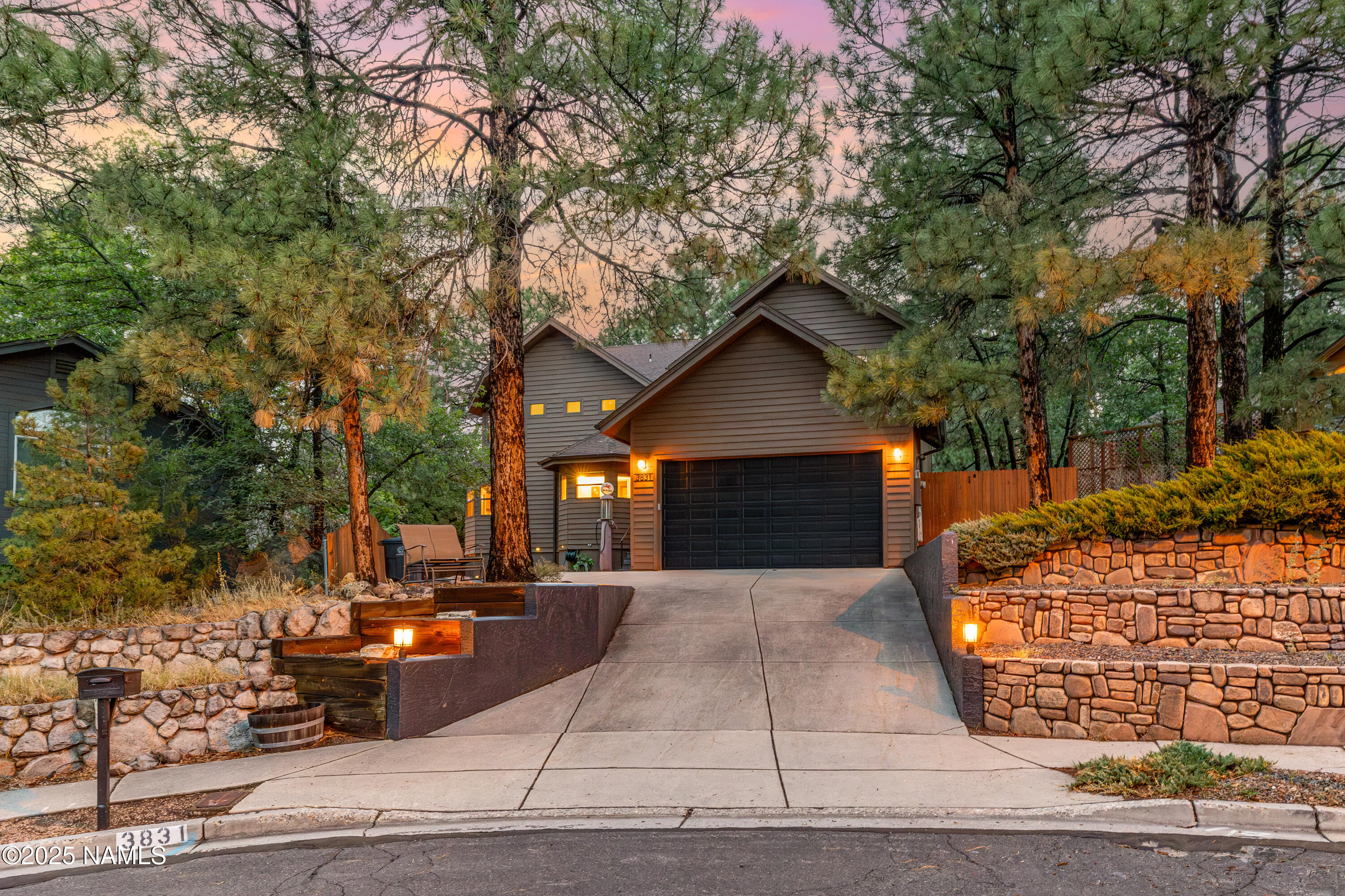 Brown two-story home with a double garage, framed by tall pines and illuminated stone landscaping at dusk.