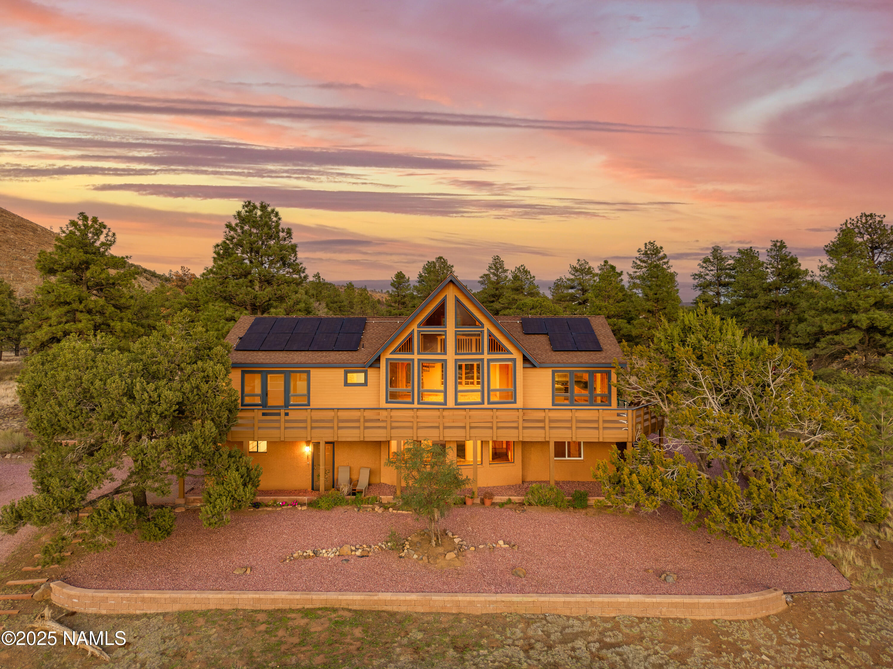 A-frame house with rooftop solar panels and a broad deck, surrounded by trees at sunset.