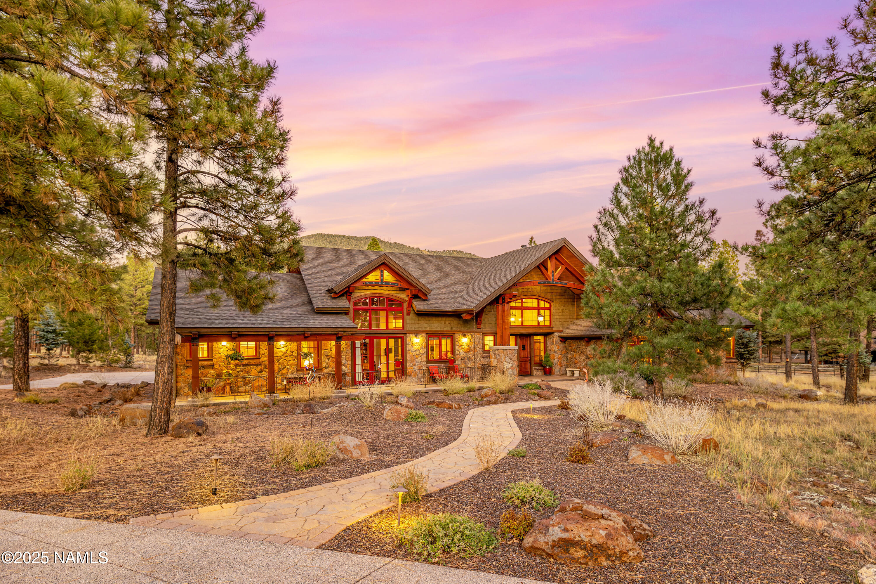 Stone-and-wood home among pines at dusk, with glowing windows and a curved stone walkway.