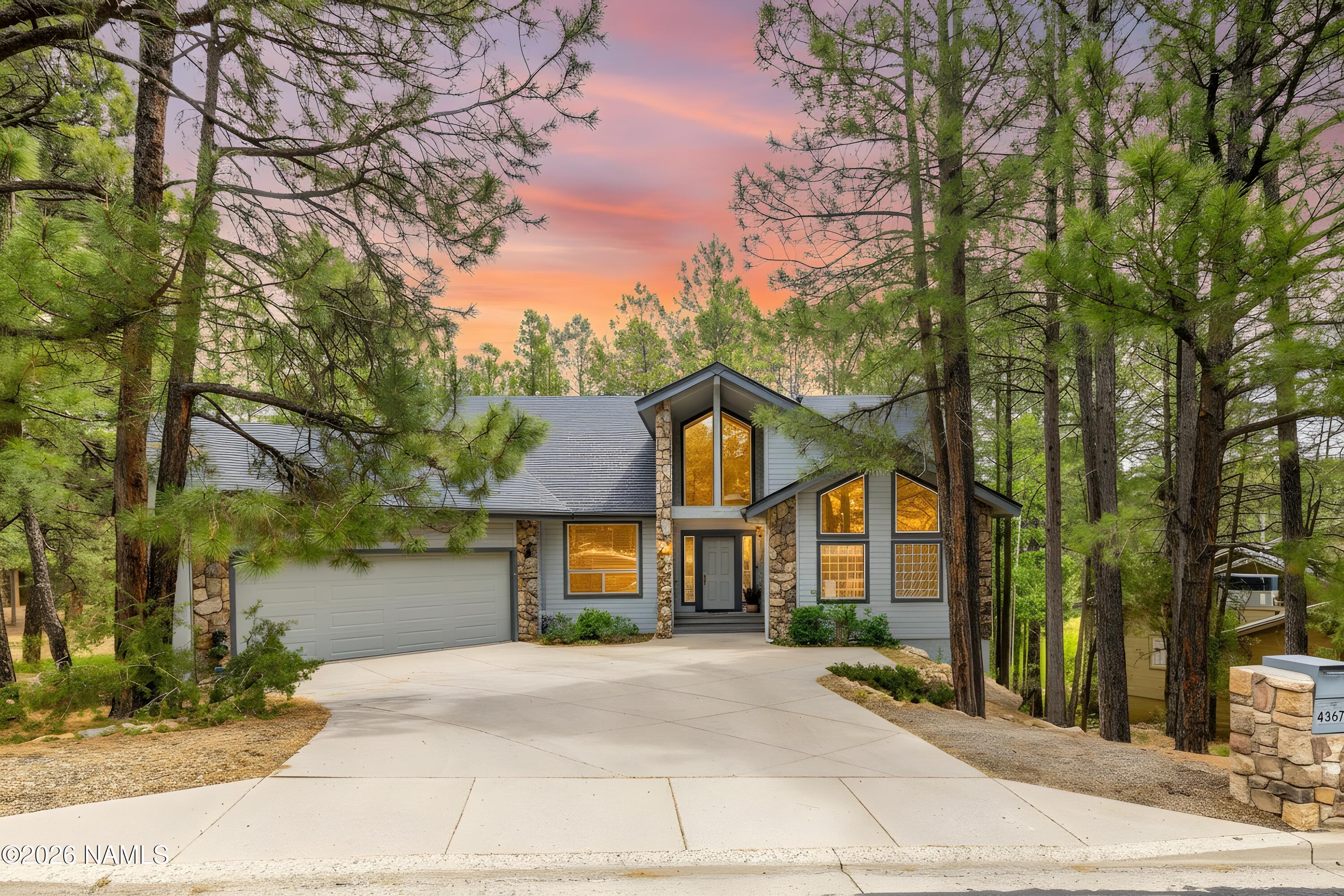 Gray two-story home with stone accents and glowing windows, surrounded by pine trees at dusk.