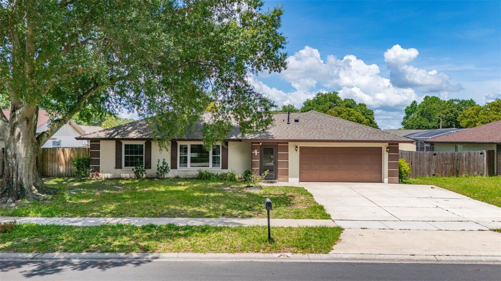 Single-story tan home with a two-car garage, broad driveway, and large shade tree in the front yard.