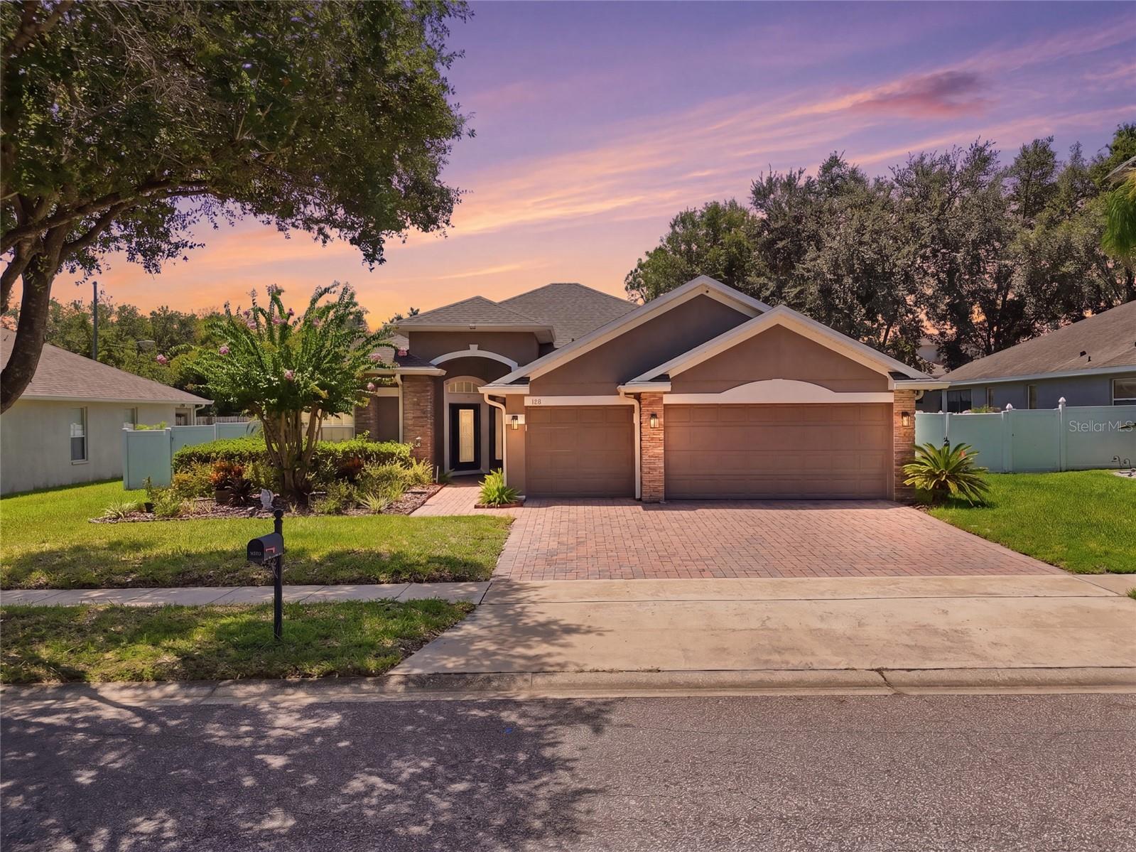 Suburban home with a three-car garage and brick driveway beneath a pink dusk sky.