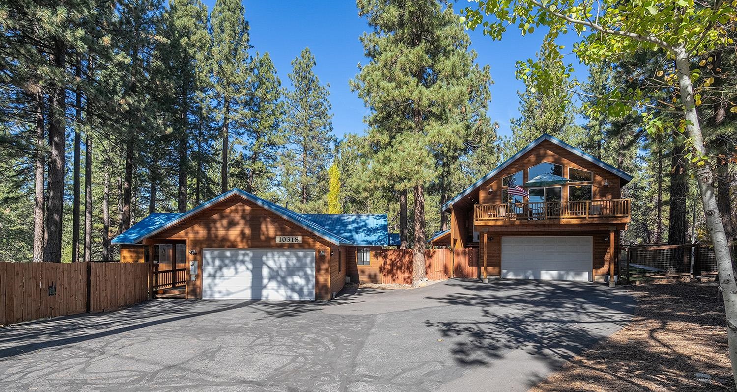Two wood-sided homes with blue roofs and garages stand among tall pine trees.