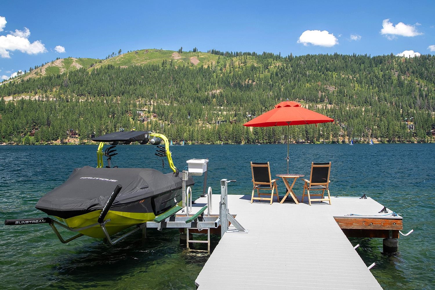 Lakeside dock with covered boat, red umbrella, chairs, and forested mountain across the water.
