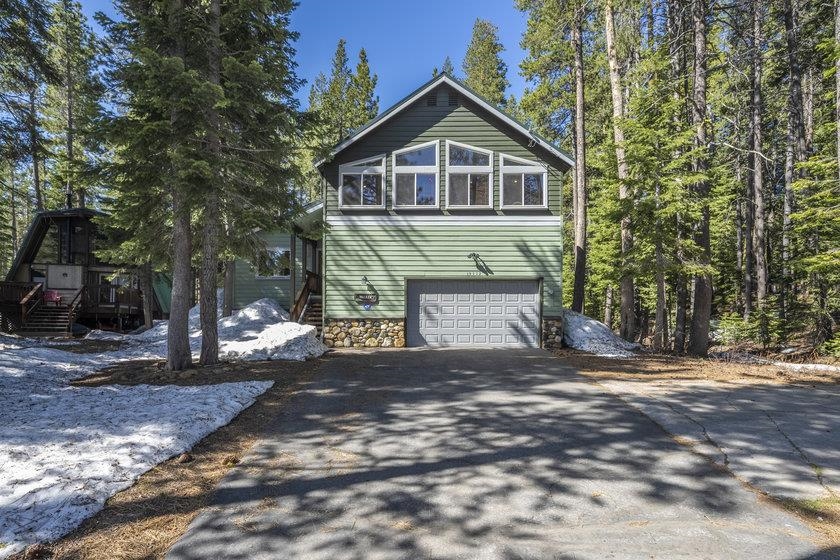 Green multi-level house with a two-car garage among evergreens, with melting snow beside the driveway.