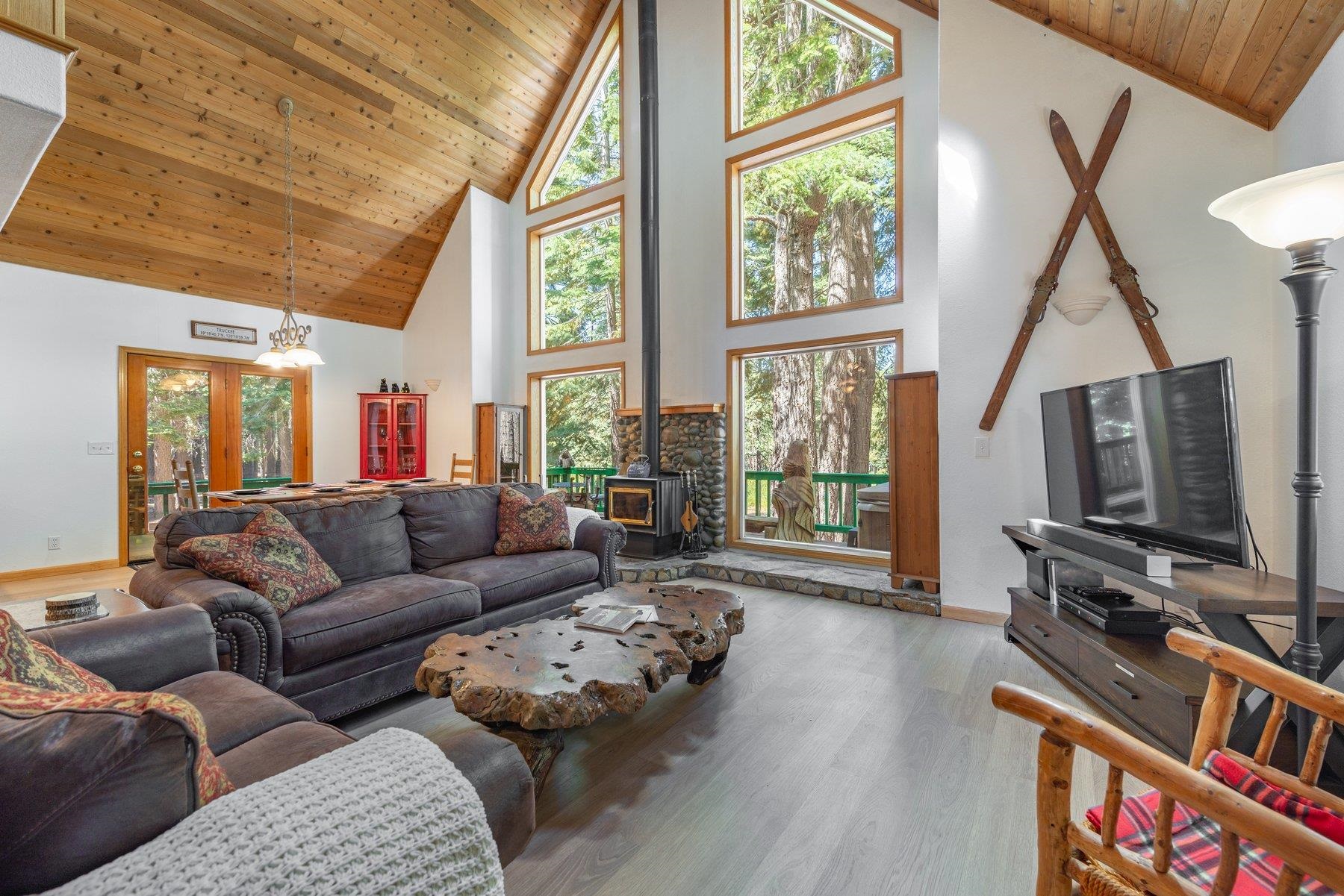 Living room with a vaulted wood ceiling, tall forest-view windows, and a stone fireplace.