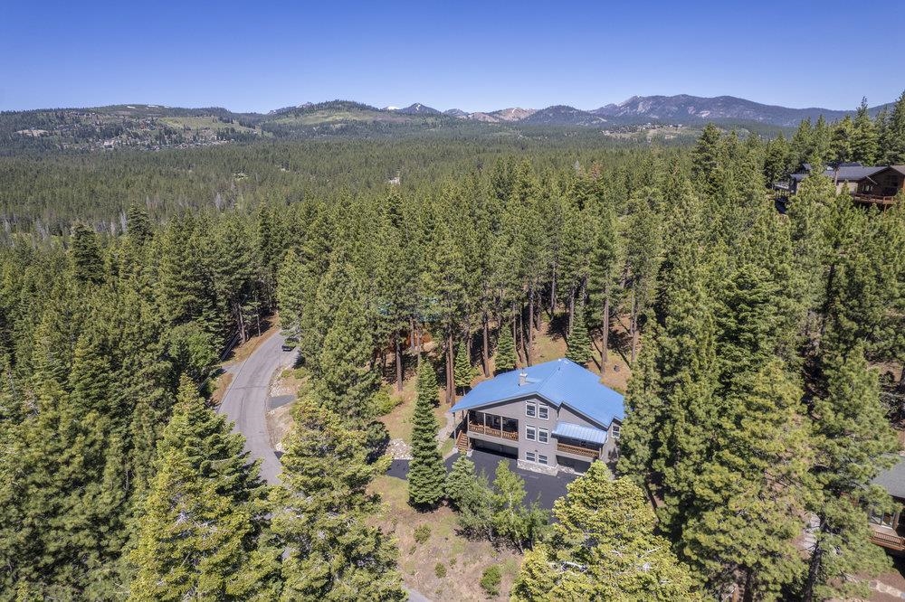 Aerial view of gray house with blue roof among pines, with distant mountains.