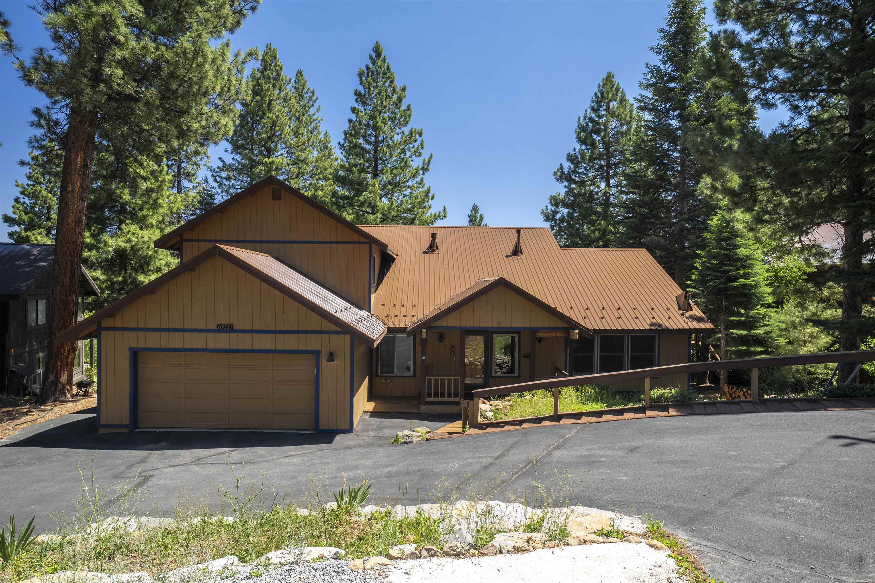 Brown mountain home with a metal roof, attached garage, and access ramp among tall pine trees.
