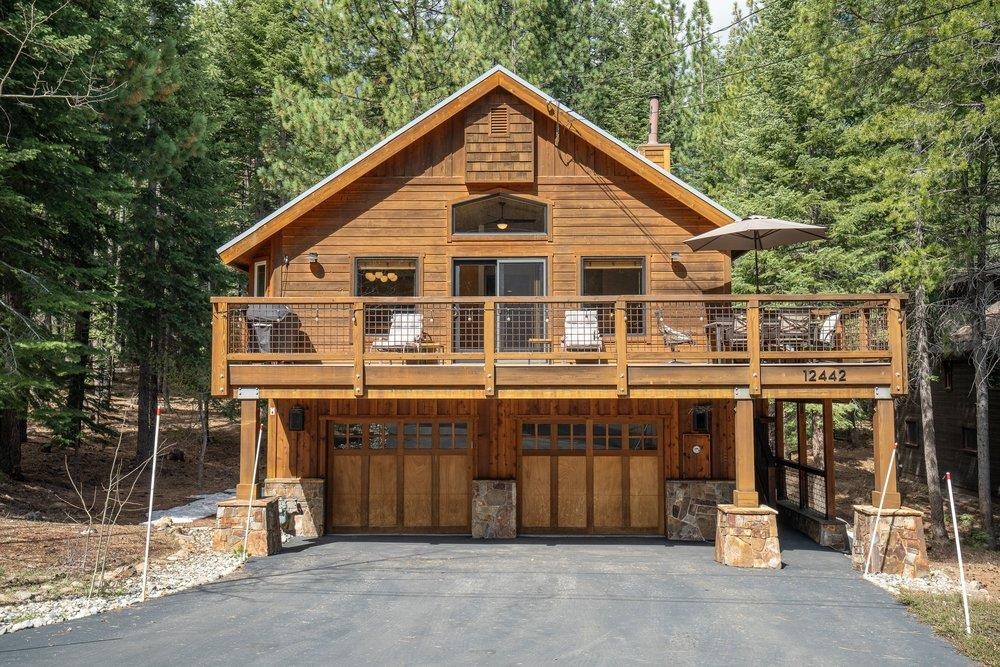 Wood cabin home with a broad deck, two garage doors, and evergreen forest surroundings.