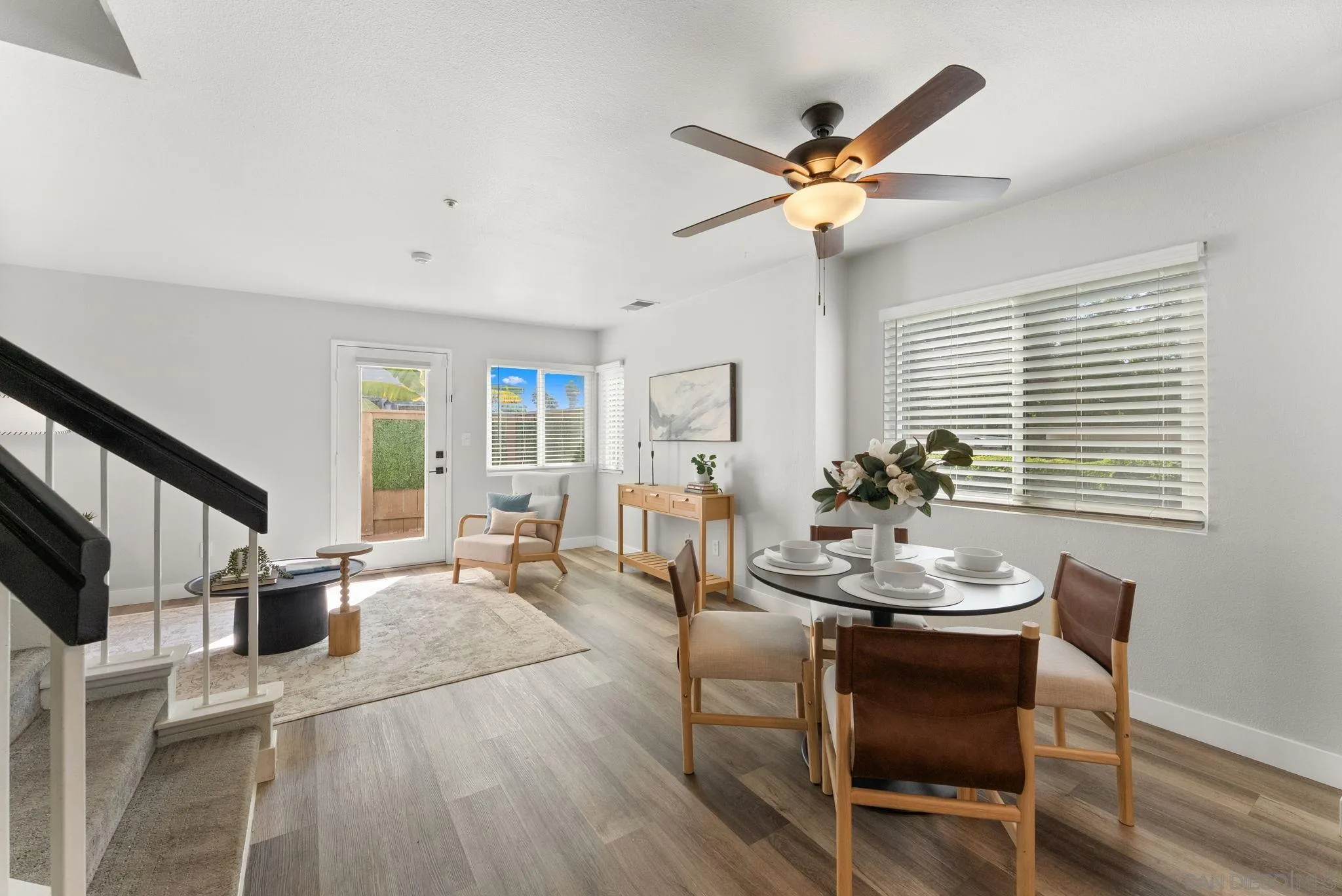 Staged dining and living area with a round table, bright windows, and a dark-railed staircase.