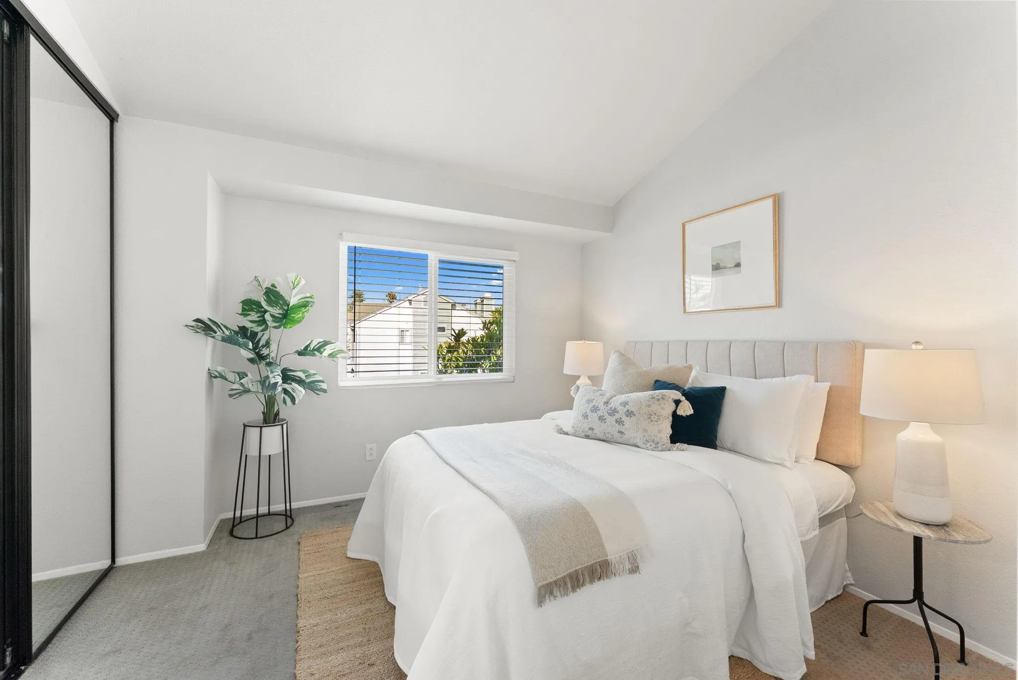 Staged bedroom with a white bed, mirrored closet doors, and a window overlooking a neighboring home.
