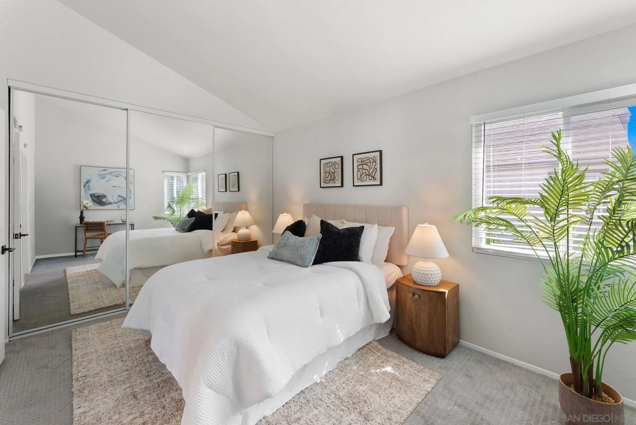 Staged bedroom with a white bed, mirrored closet doors, and a large window beside a potted palm.