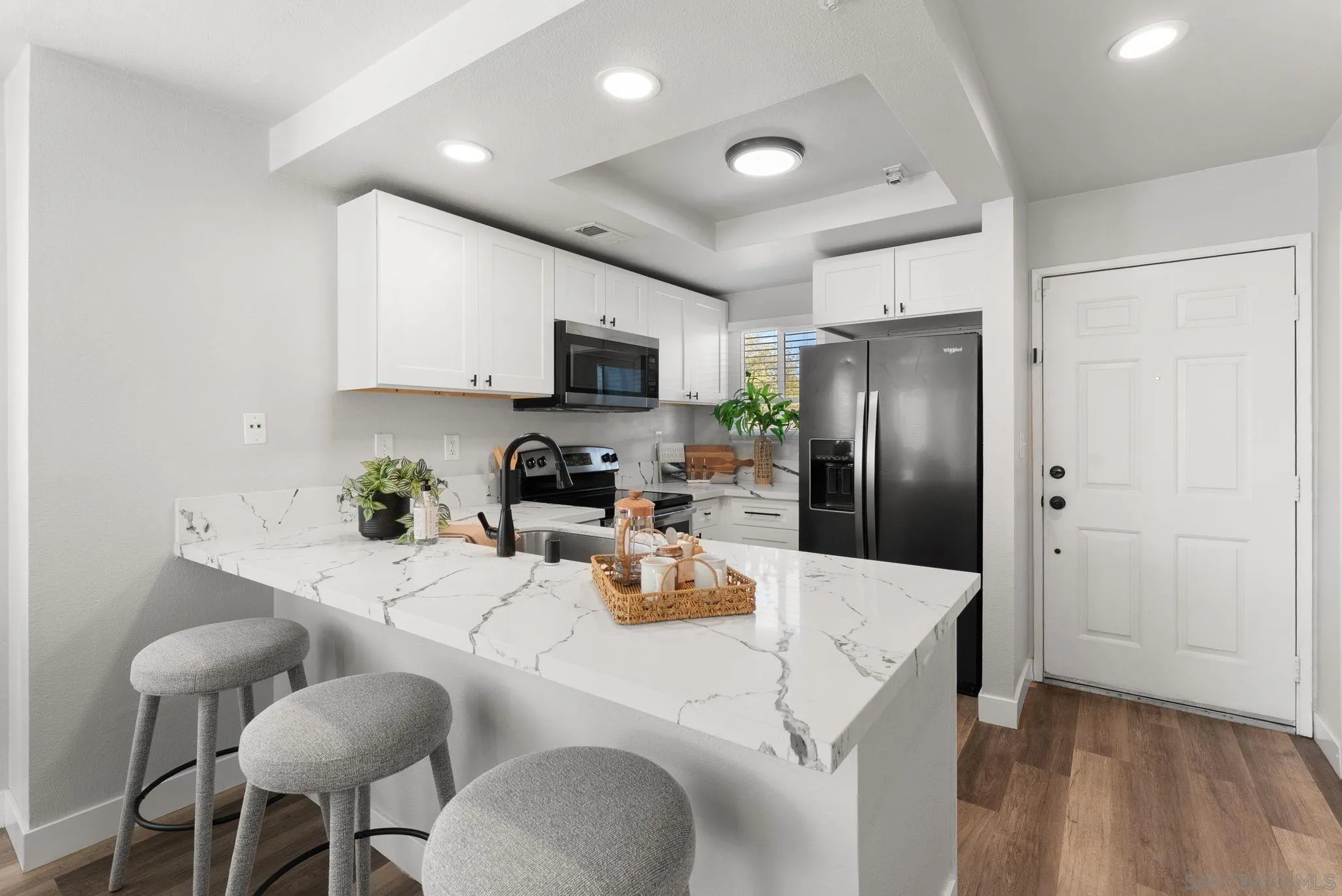 Kitchen with a veined white island, three gray stools, white cabinets, and black appliances.