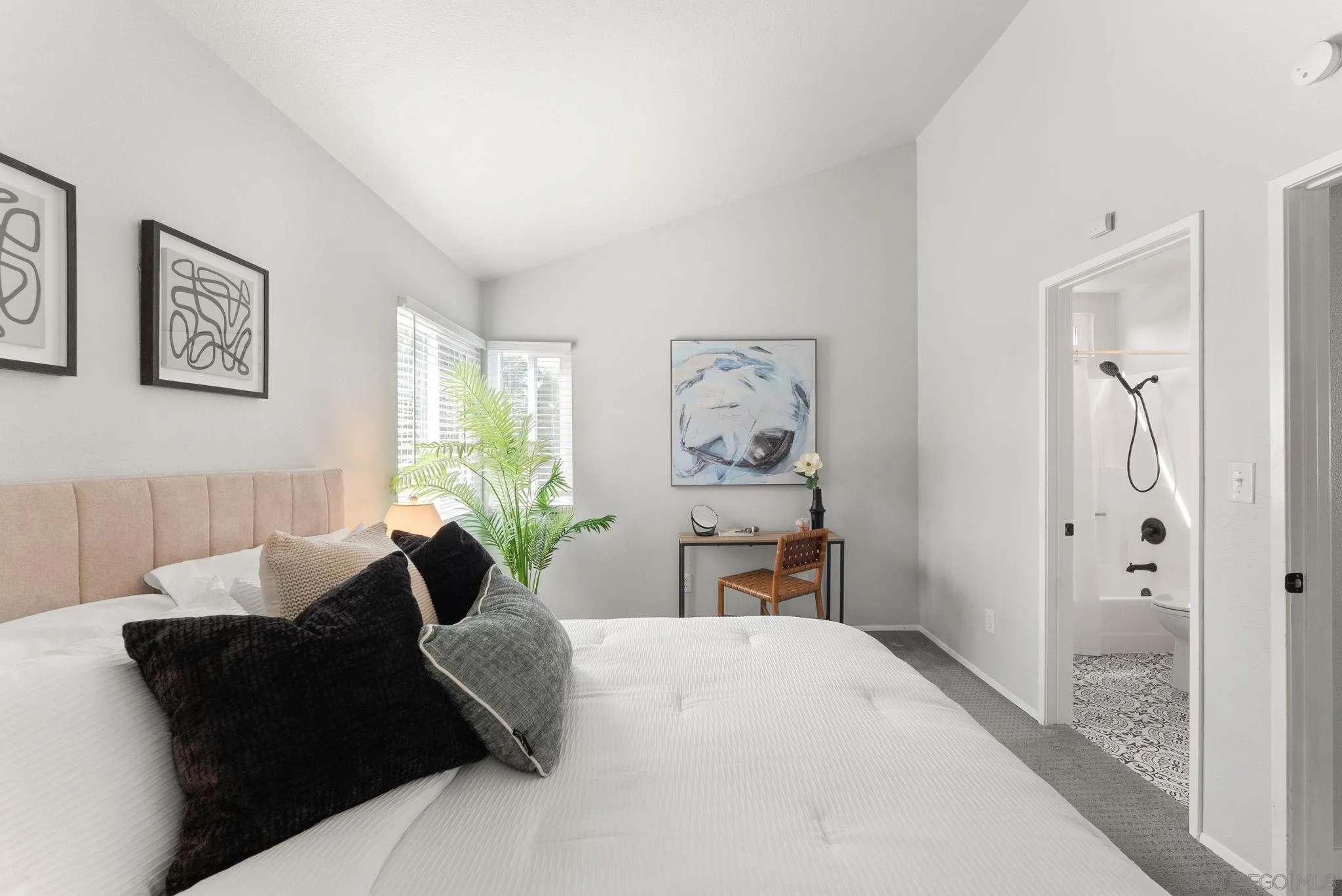 Staged bedroom with a white bed, blush headboard, potted palm, and adjoining bathroom.
