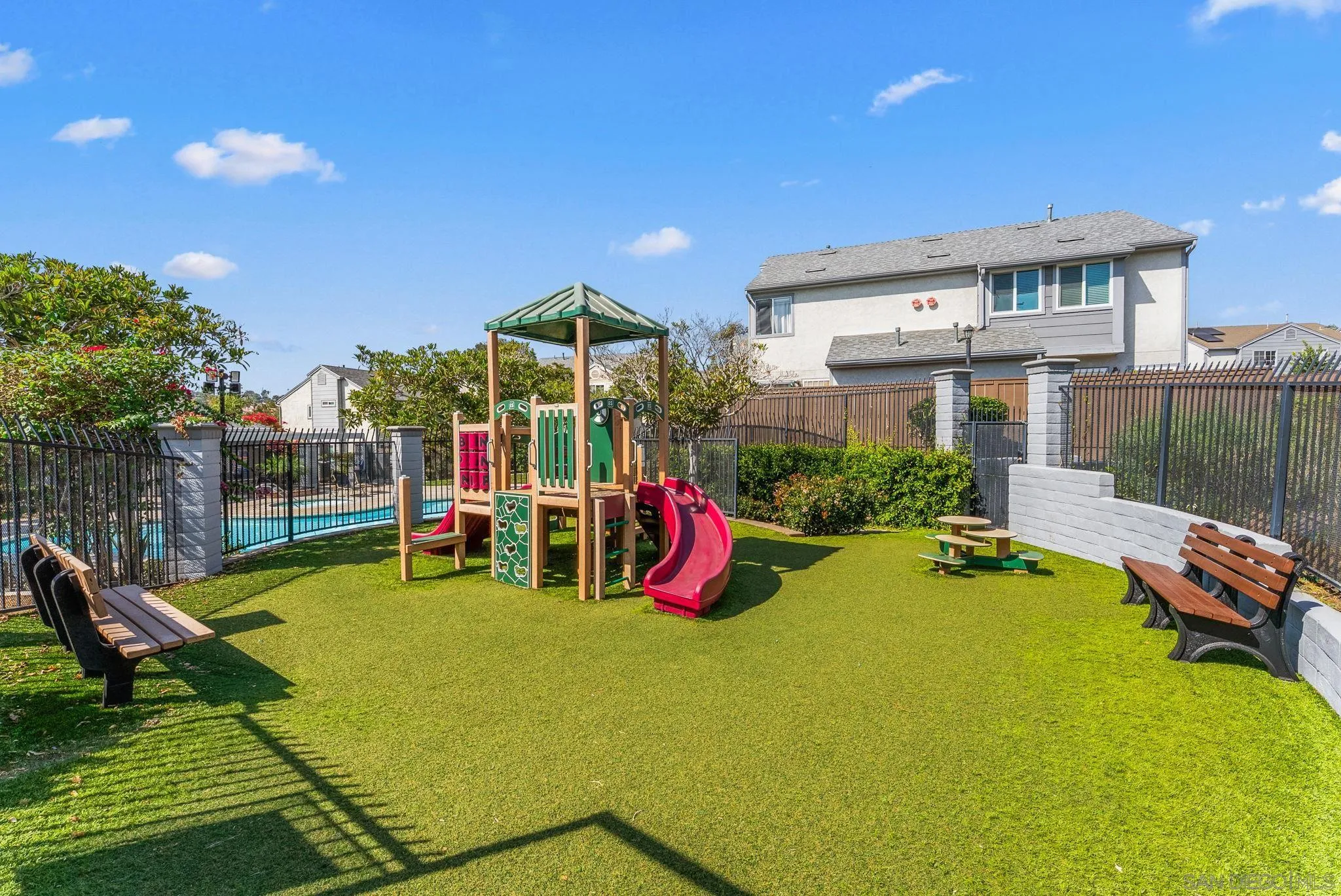 Fenced outdoor playground with a colorful play structure, artificial turf, benches, and a swimming pool.