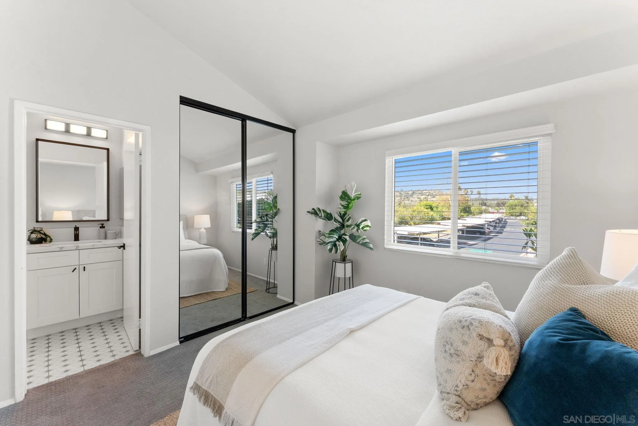 Bright bedroom with a white bed, mirrored closet doors, and an adjoining bathroom vanity.