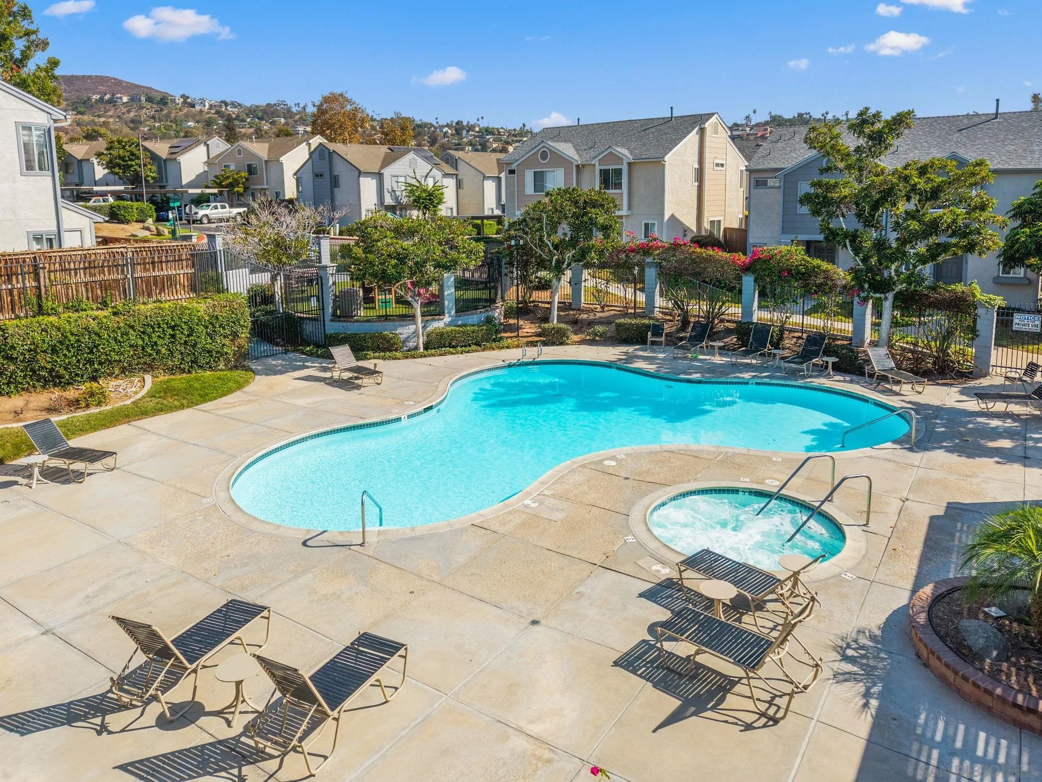 Community pool and bubbling spa surrounded by lounge chairs, landscaping, and nearby homes.