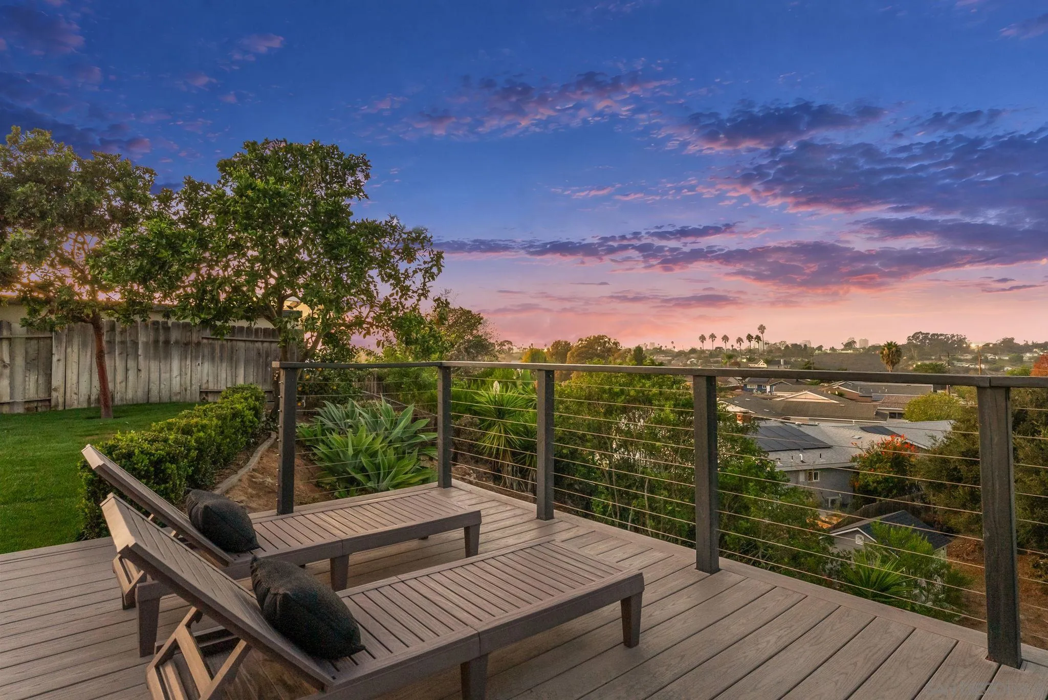 Deck with lounge chairs and cable railing overlooking neighborhood rooftops at dusk.