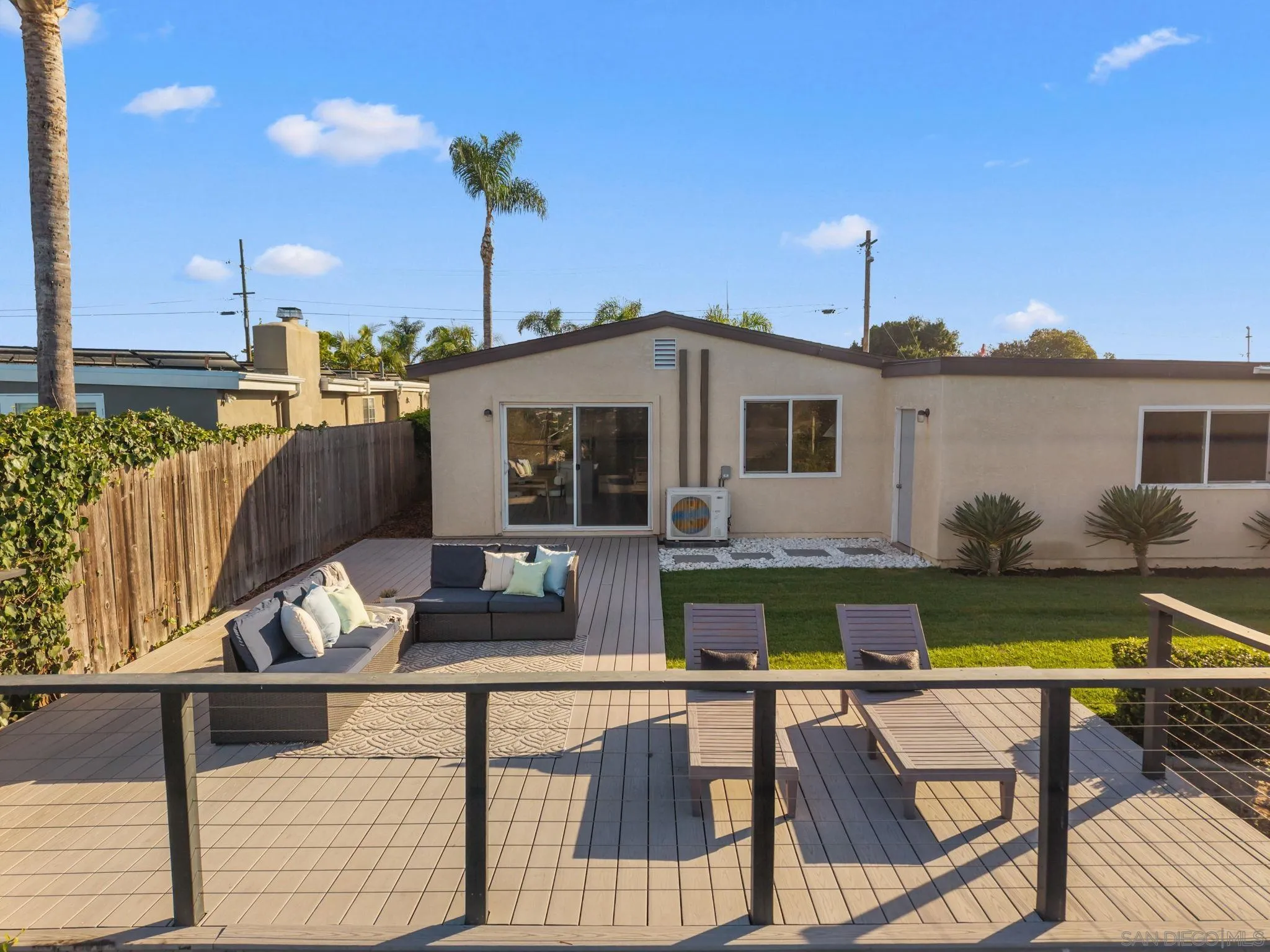 Home’s rear exterior with a furnished raised deck, lawn, and palm trees.