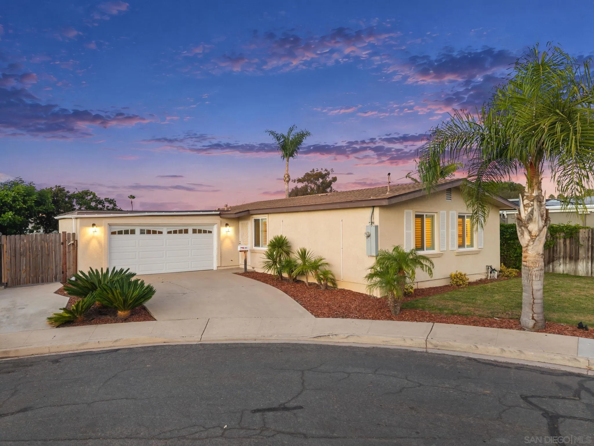 Single-story beige home with an attached two-car garage, palm landscaping, and illuminated windows at dusk.