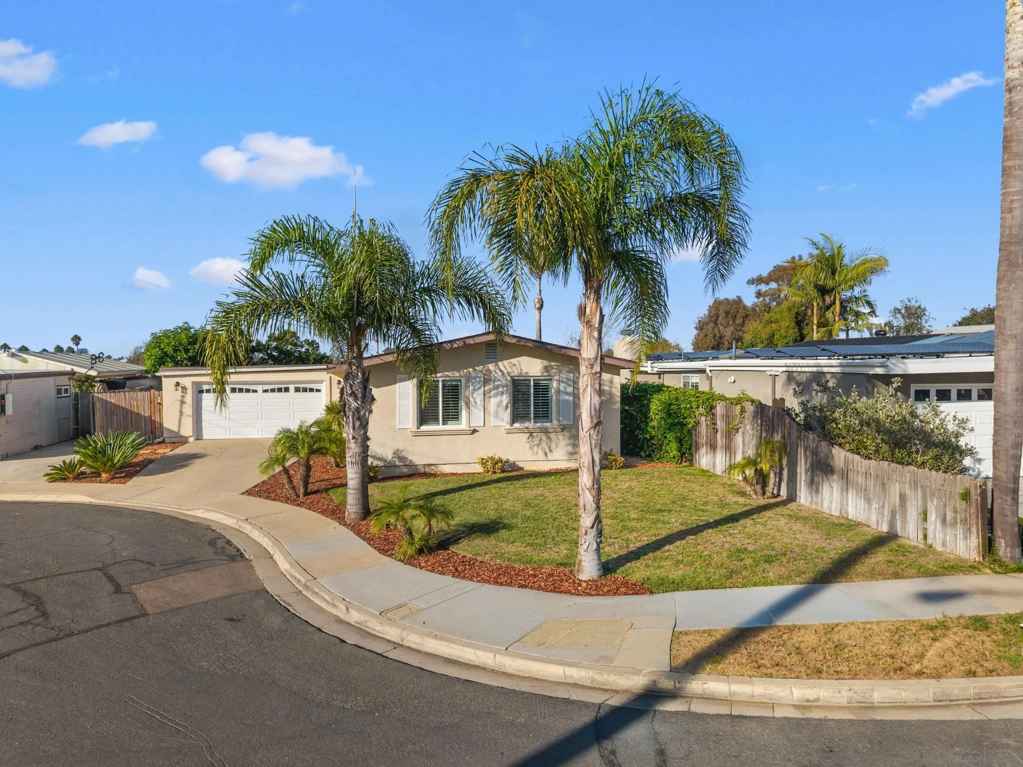 Single-story beige home with palm trees, a corner lawn, and a driveway to the garage.
