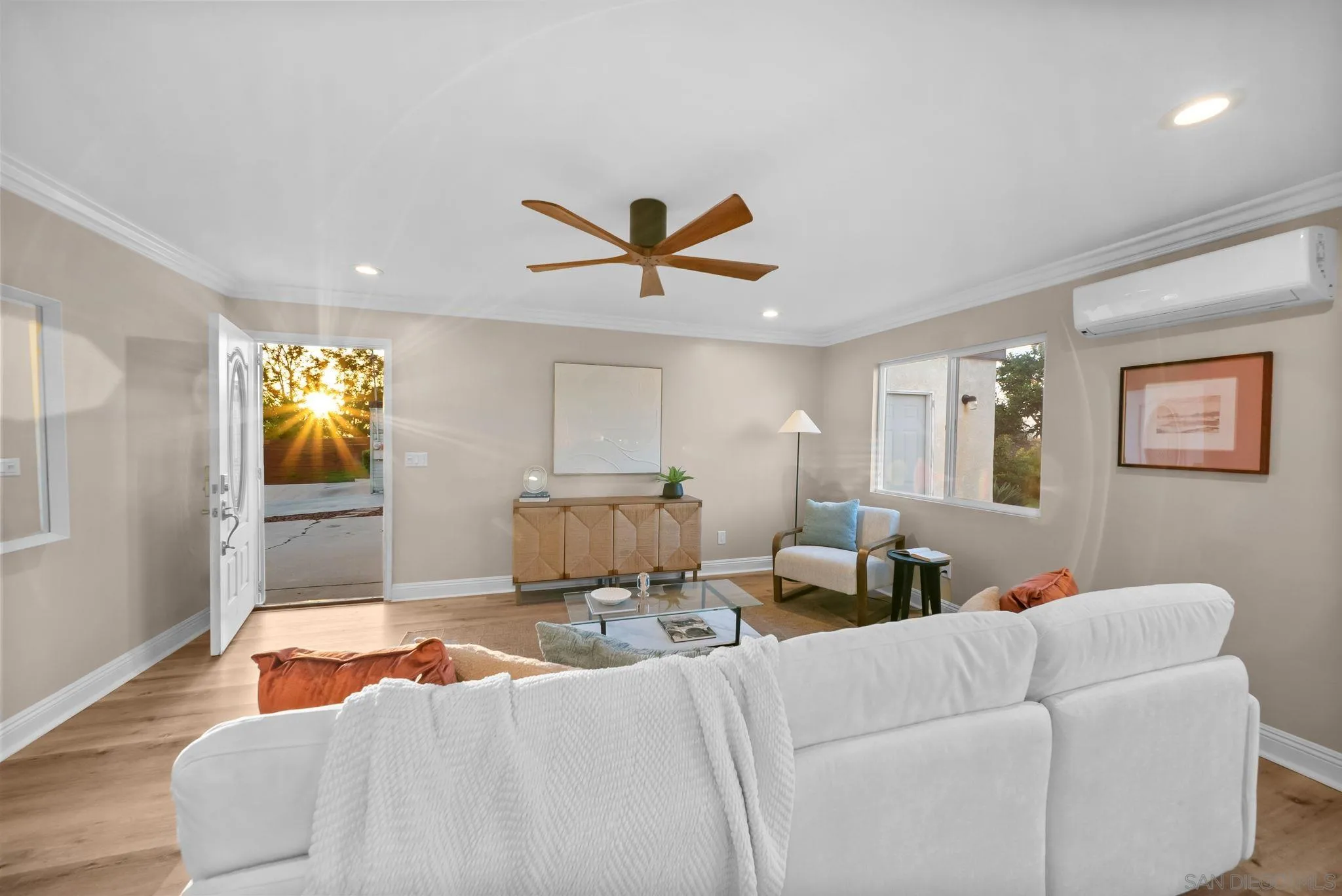 Staged living room with a white sectional, wood floors, and golden-hour sunlight streaming through the open front door.