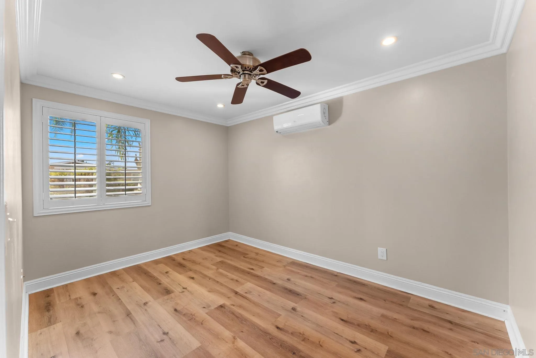 Empty bedroom with wood plank flooring, plantation shutters, ceiling fan, and wall-mounted mini-split.