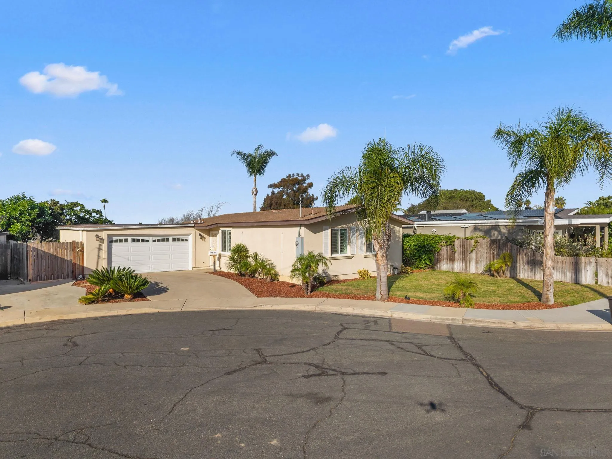 Single-story beige home with an attached two-car garage, palm trees, and a curved driveway.